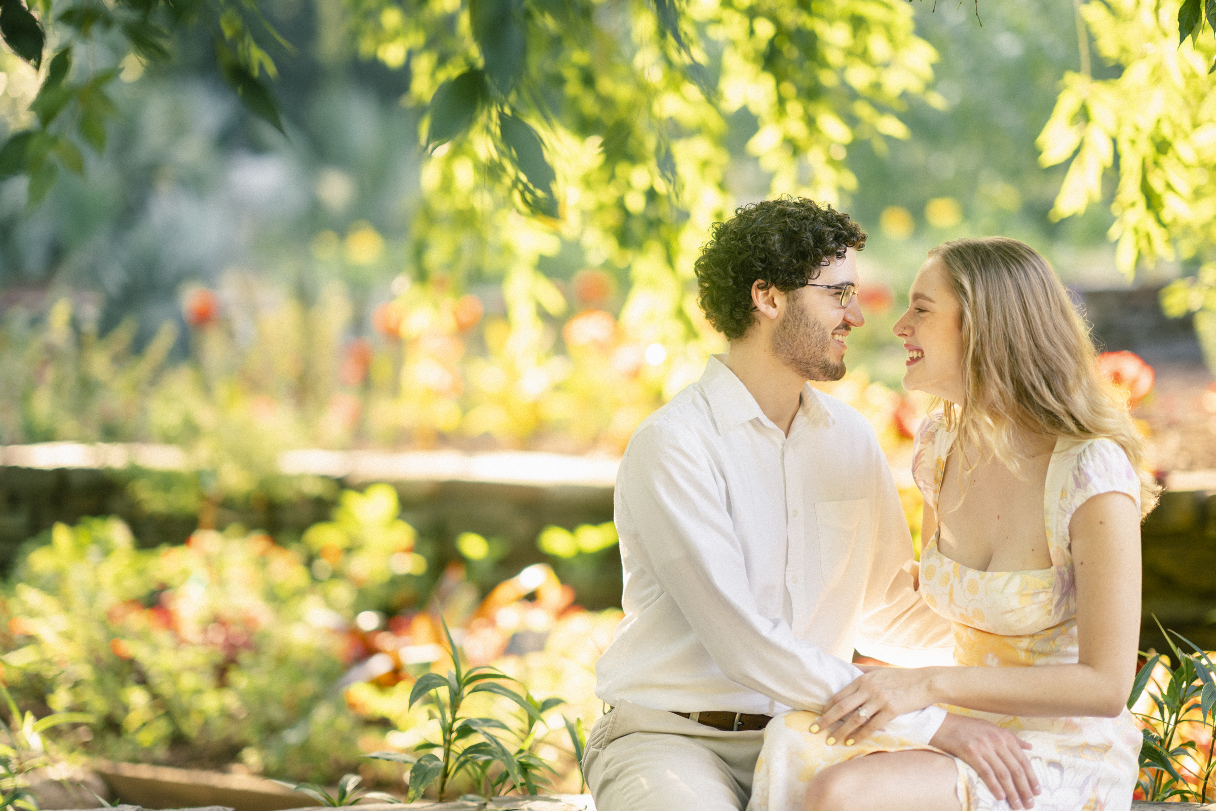 In the heart of Duke Gardens, a couple sits closely on a stone bench, basking in the sunlight. They exchange smiles amid vibrant flowers and lush greenery, creating a warm and serene atmosphere perfect for their engagement session.