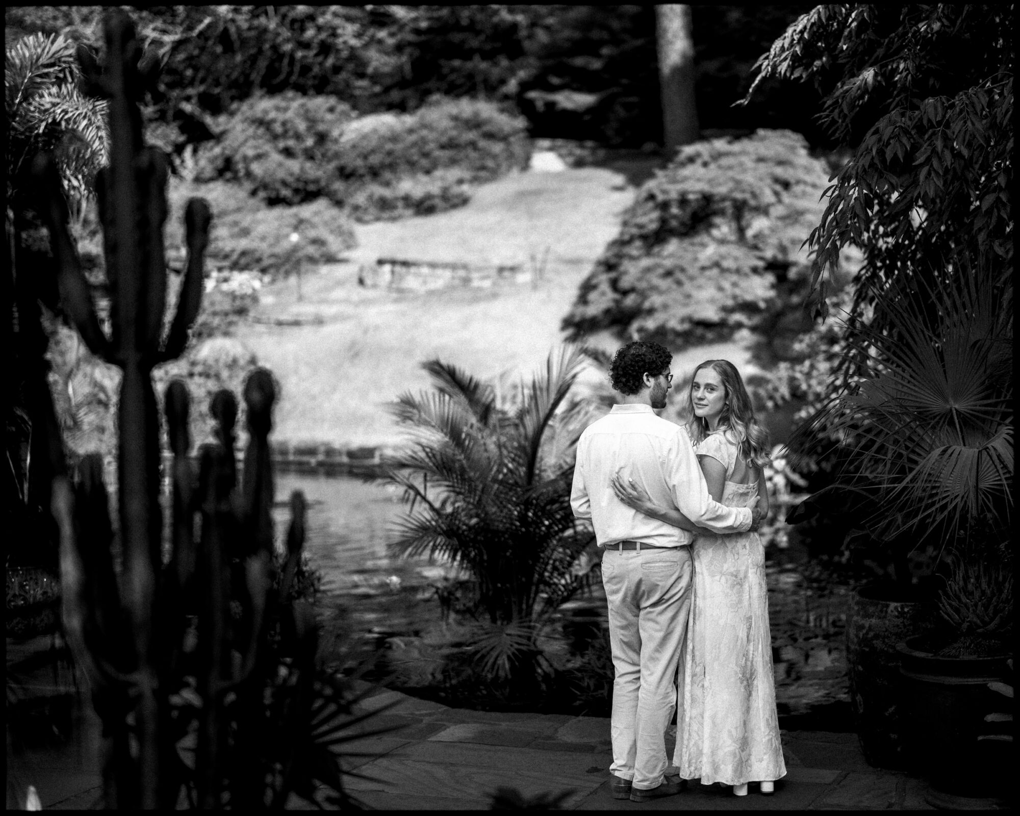 A couple stands together outdoors near a pond, surrounded by the lush greenery and cacti of Duke Gardens. The woman, wearing a flowing dress, looks over her shoulder as the man, in a light shirt and pants, faces her. The scene exudes a serene, romantic atmosphere perfect for an engagement session.