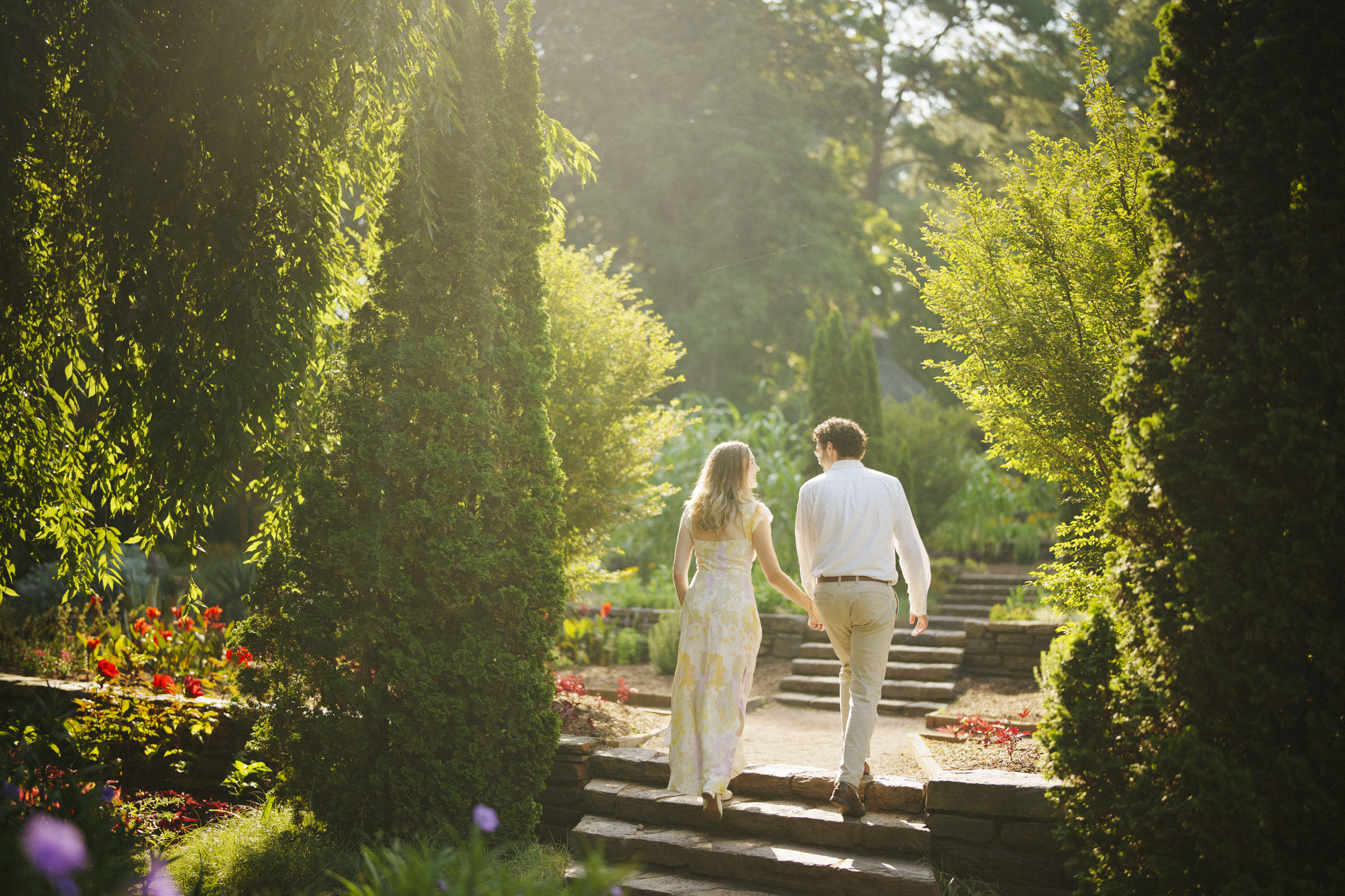 A couple walks hand in hand up a stone path surrounded by lush greenery and sunlight filtering through the trees in a serene Duke Gardens engagement session.