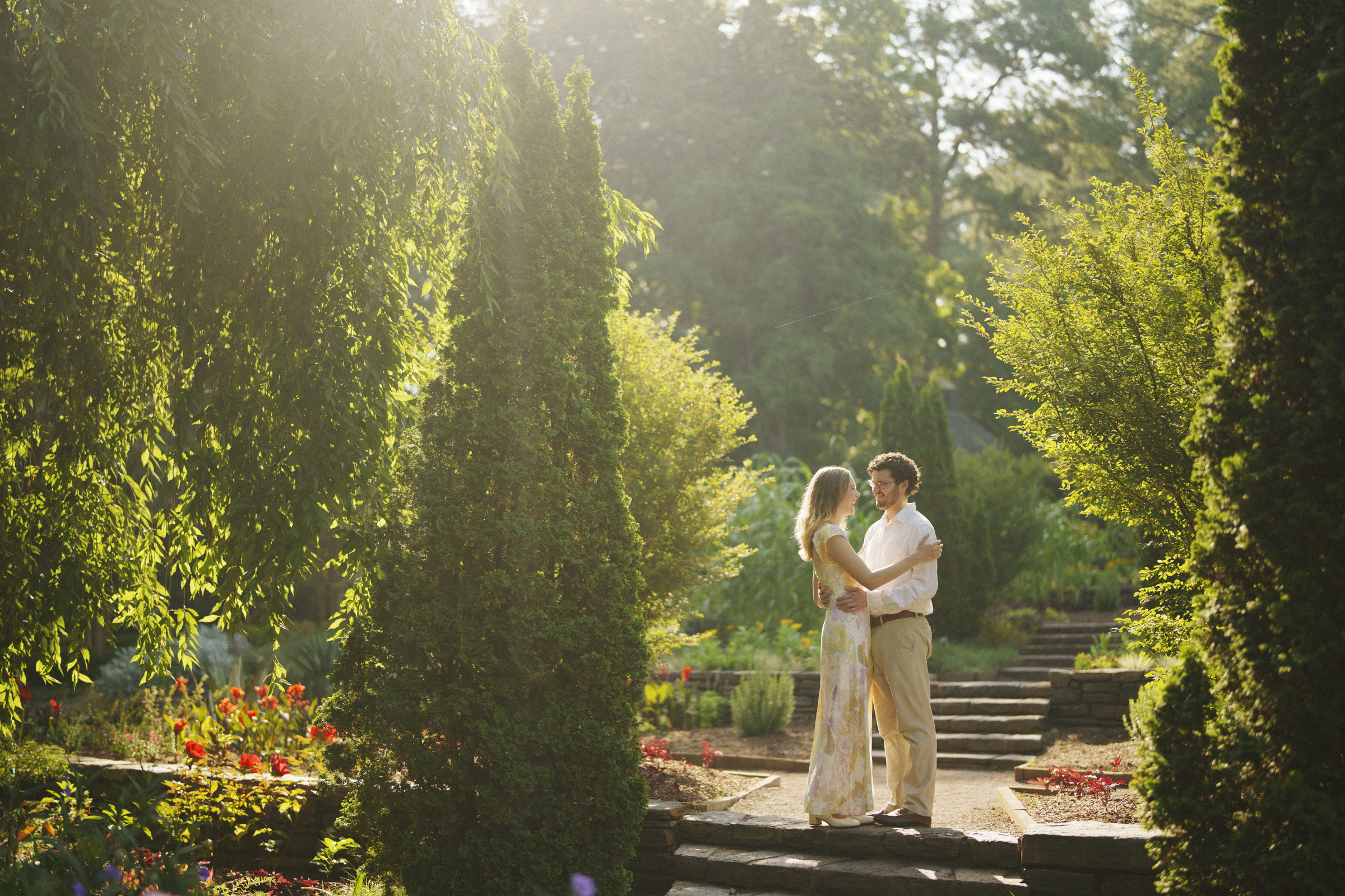 A couple stands on a stone pathway in Duke Gardens, surrounded by tall, sunlit trees and vibrant flowers, warmly gazing at each other. The sunlight filters through the foliage, creating a serene, romantic atmosphere for their engagement session.