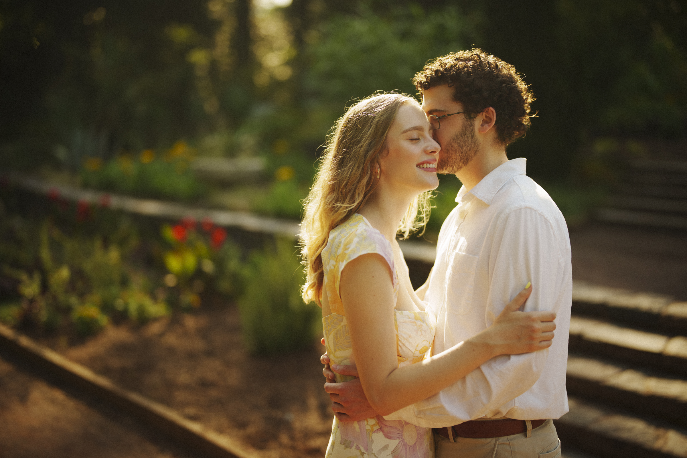 The couple embraces in a sunlit garden, surrounded by lush greenery and colorful flowers during their Duke Gardens engagement session. The woman, in a floral dress, smiles as the man, wearing a white shirt, gently leans in for a tender moment.