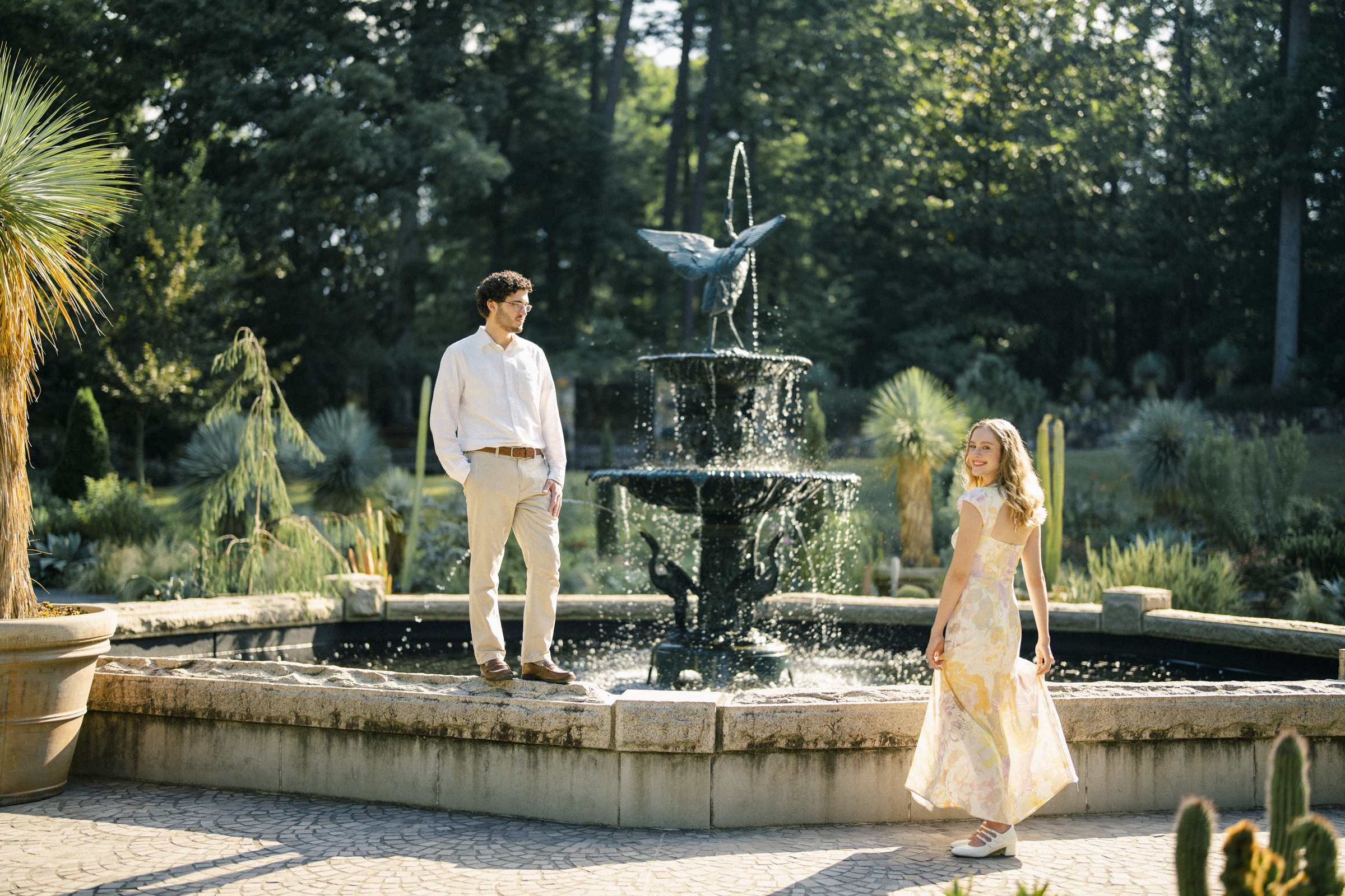 In the midst of Duke Gardens, a man and woman share a serene engagement session. The man stands on the fountain's edge in a white shirt and beige pants, while the woman in a floral dress strolls nearby. They are enveloped by lush greenery under bright sunlight, capturing a moment of bliss.