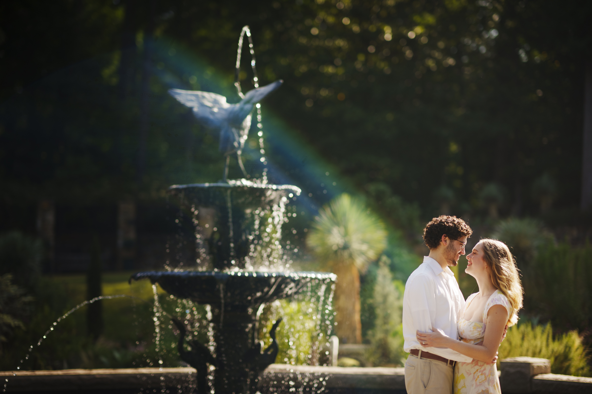 In Duke Gardens, during an enchanting engagement session, a couple embraces in front of a fountain with water cascading from multiple tiers. Sunlight creates a rainbow effect in the mist as they stand near lush greenery, gazing at each other affectionately.