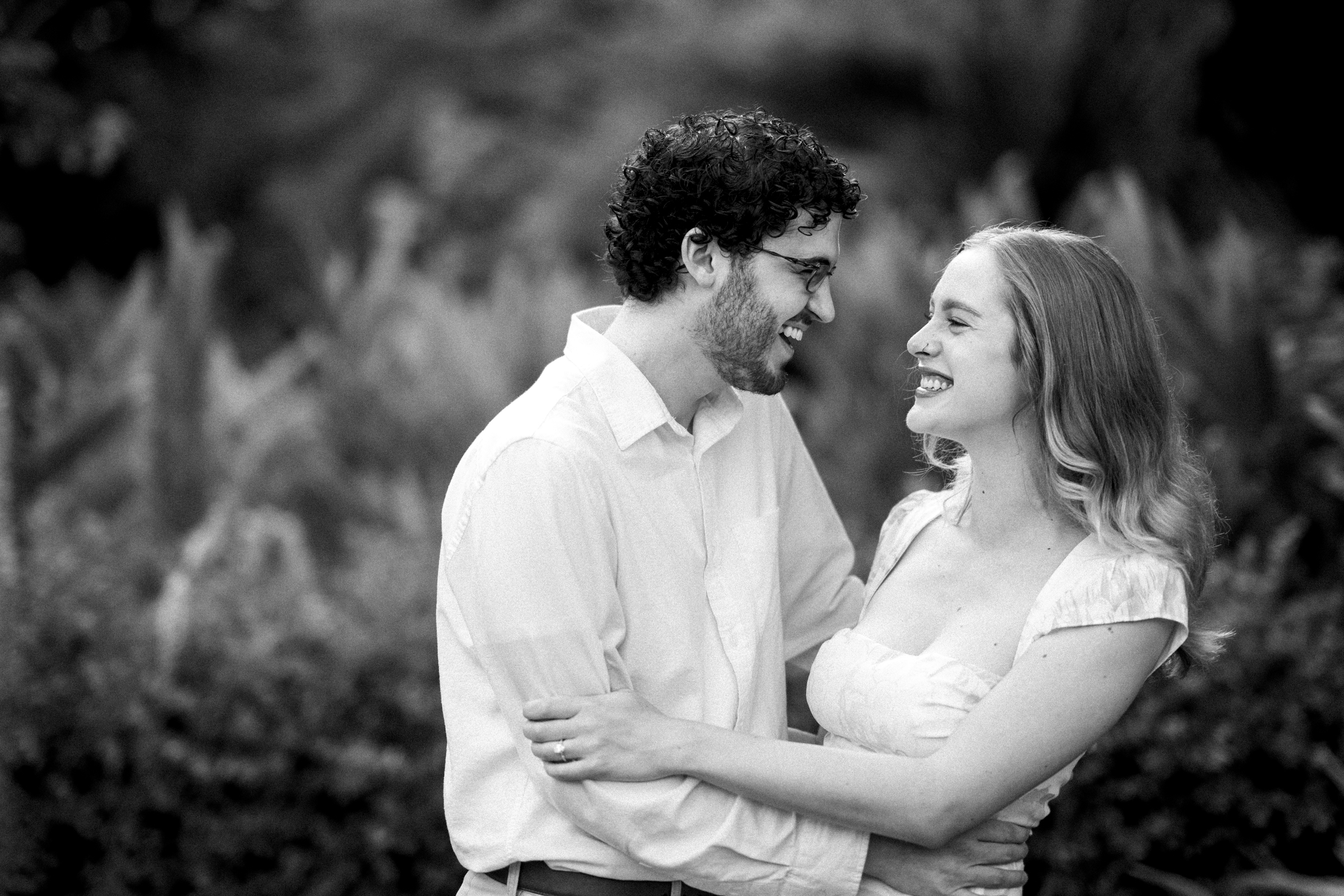 A black and white photo captures a couple laughing and looking at each other during a Duke Gardens engagement session. The man with curly hair and glasses wears a light shirt, while the woman with long hair dons a light dress. They embrace outdoors against a blurred leafy backdrop.