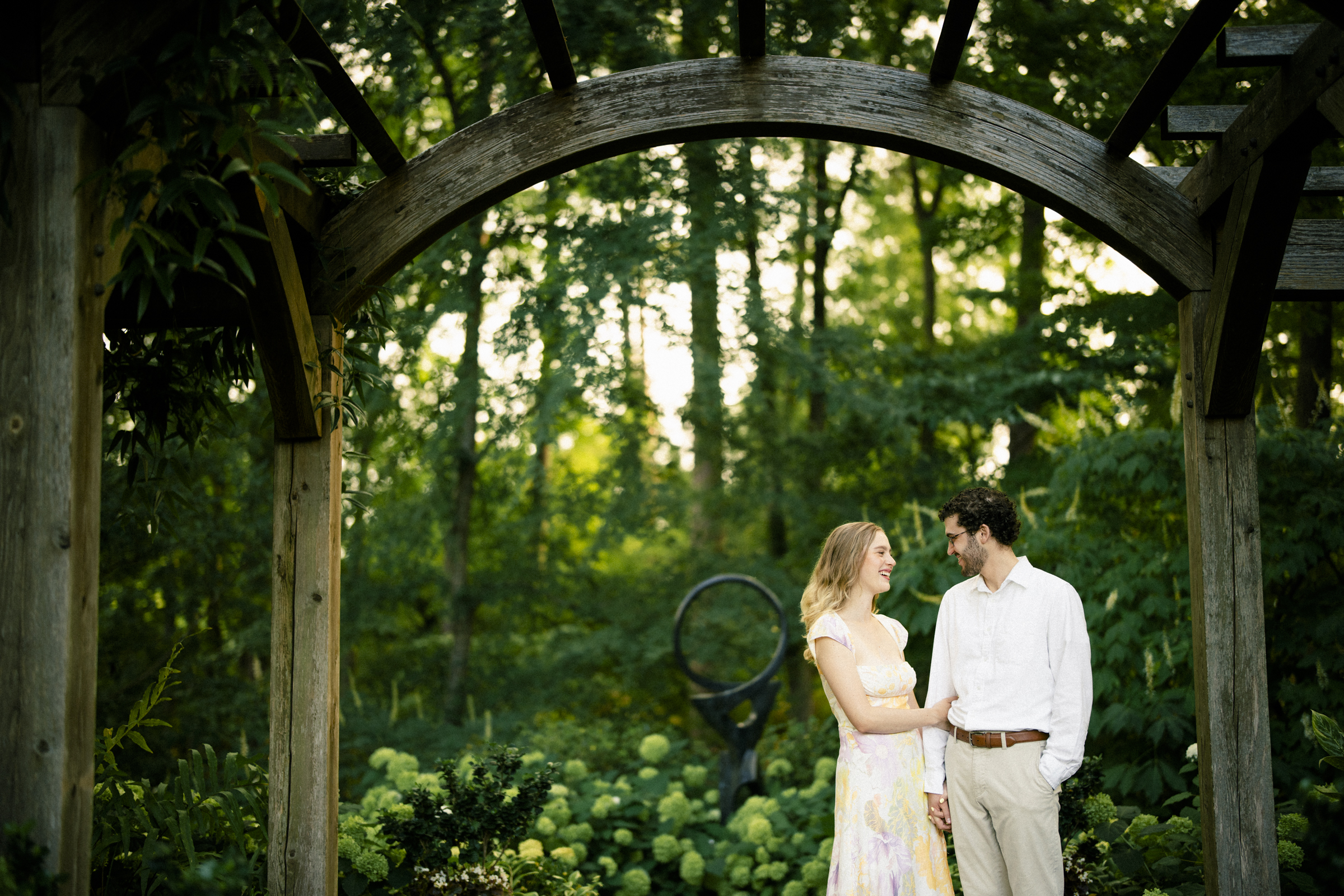 In a romantic and serene Duke Gardens engagement session, a couple stands under a wooden arbor, surrounded by lush greenery and blooming hydrangeas. Smiling at each other while holding hands, they perfectly encapsulate the beauty of the moment.