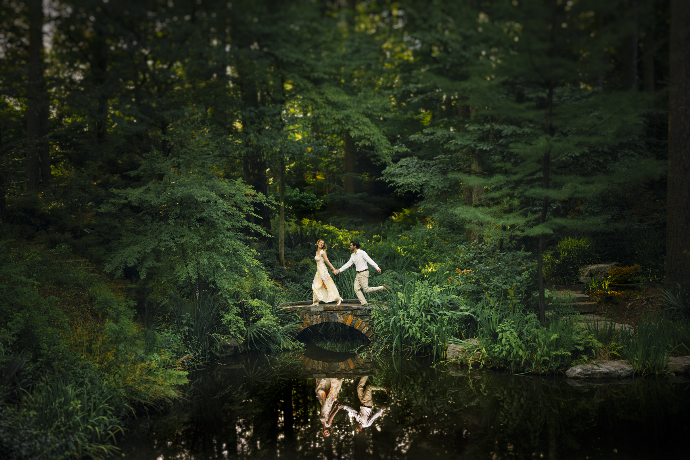 In a scene reminiscent of a Duke Gardens engagement session, a couple in elegant attire walks hand in hand across a small stone bridge over a pond in a lush, green forest. Their reflection is visible in the water below, surrounded by thick foliage and trees.