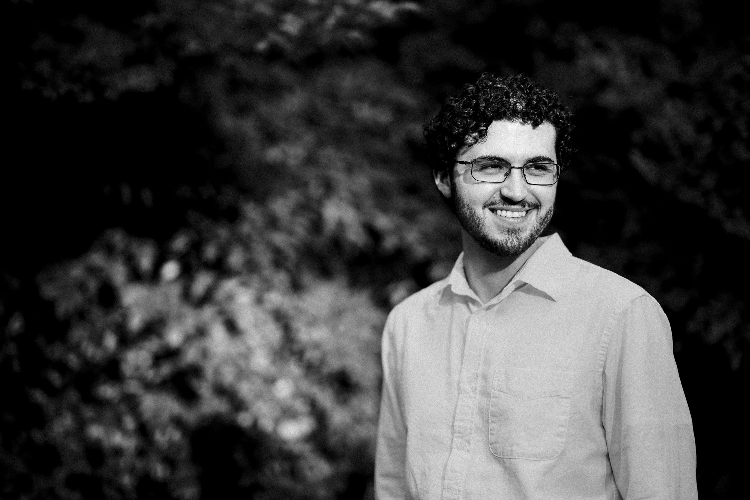 A man with curly hair and glasses smiles during an engagement session at Duke Gardens. He wears a light-colored shirt, surrounded by a blurred natural background that creates a peaceful and relaxed atmosphere. The black-and-white image captures the timeless serenity of the moment.