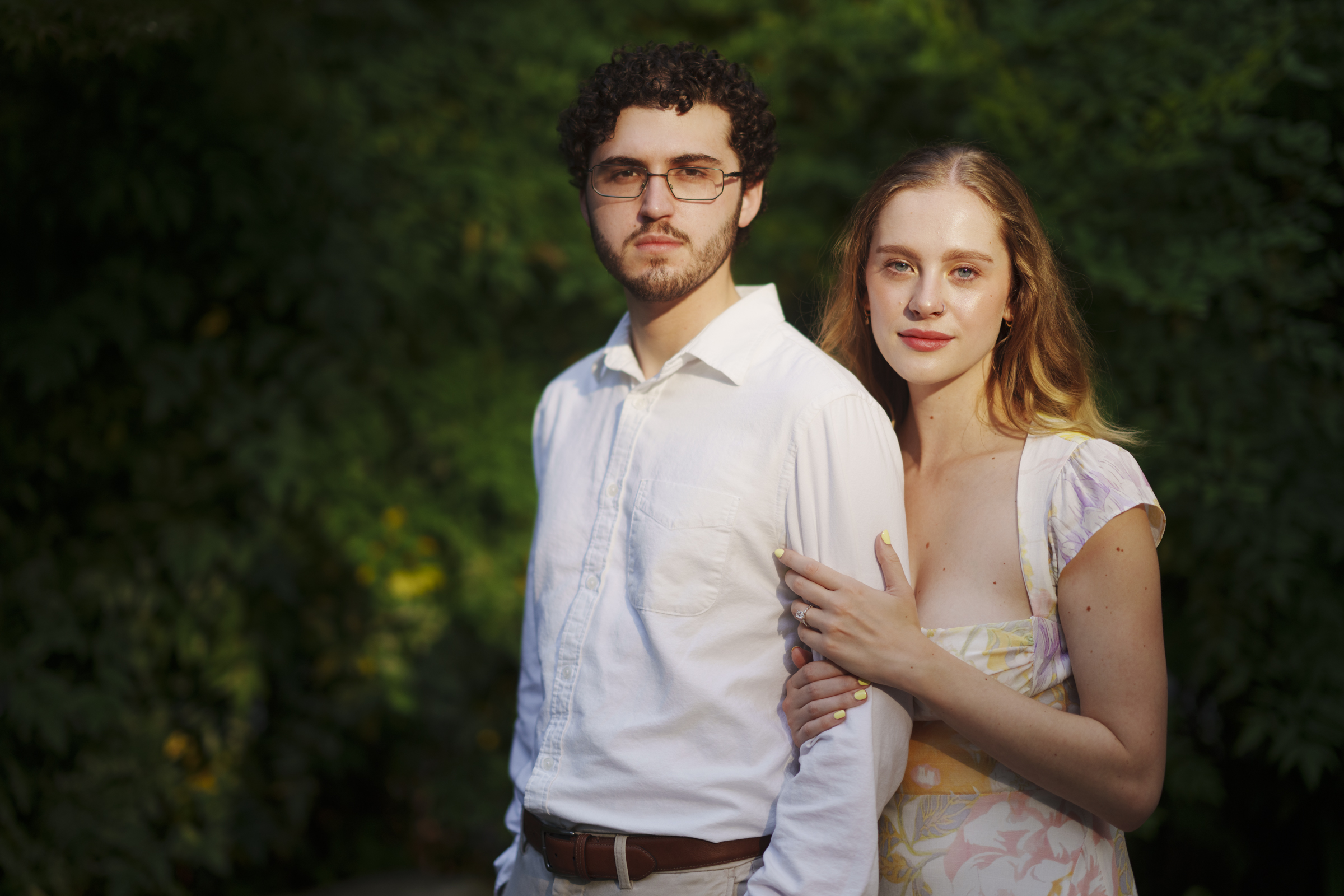 A couple stands close together in Duke Gardens, with a woman gently holding a man's arm. Surrounded by lush green foliage, the engagement session captures their joy. The man wears glasses and a white shirt, while the woman has long hair and dons a light patterned dress.