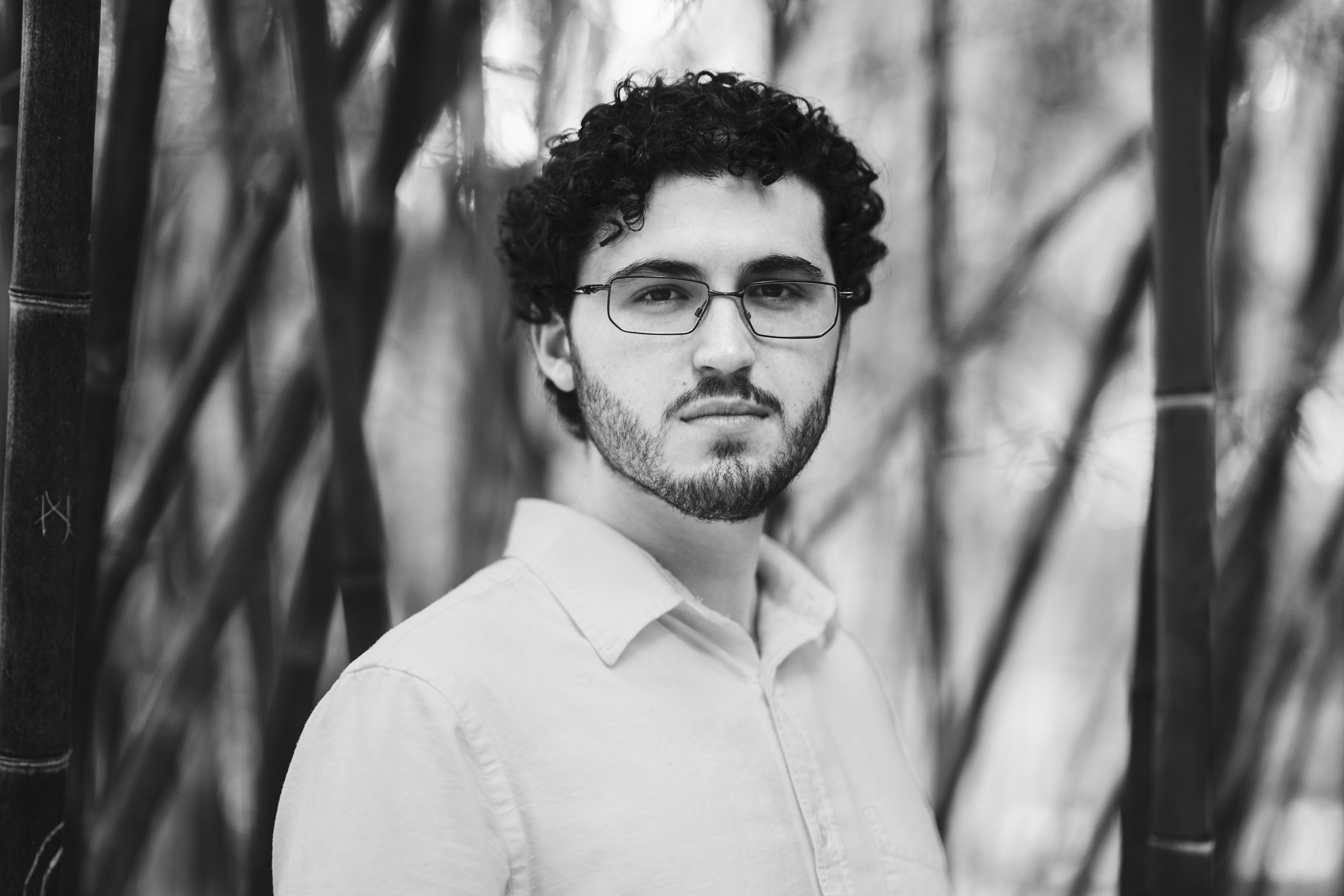 A monochrome portrait of a person with curly hair and glasses, wearing a light-colored shirt. Standing among tall bamboo stalks, reminiscent of an enchanting Duke Gardens engagement session, they gaze directly at the camera. The background is softly blurred, highlighting their thoughtful expression.