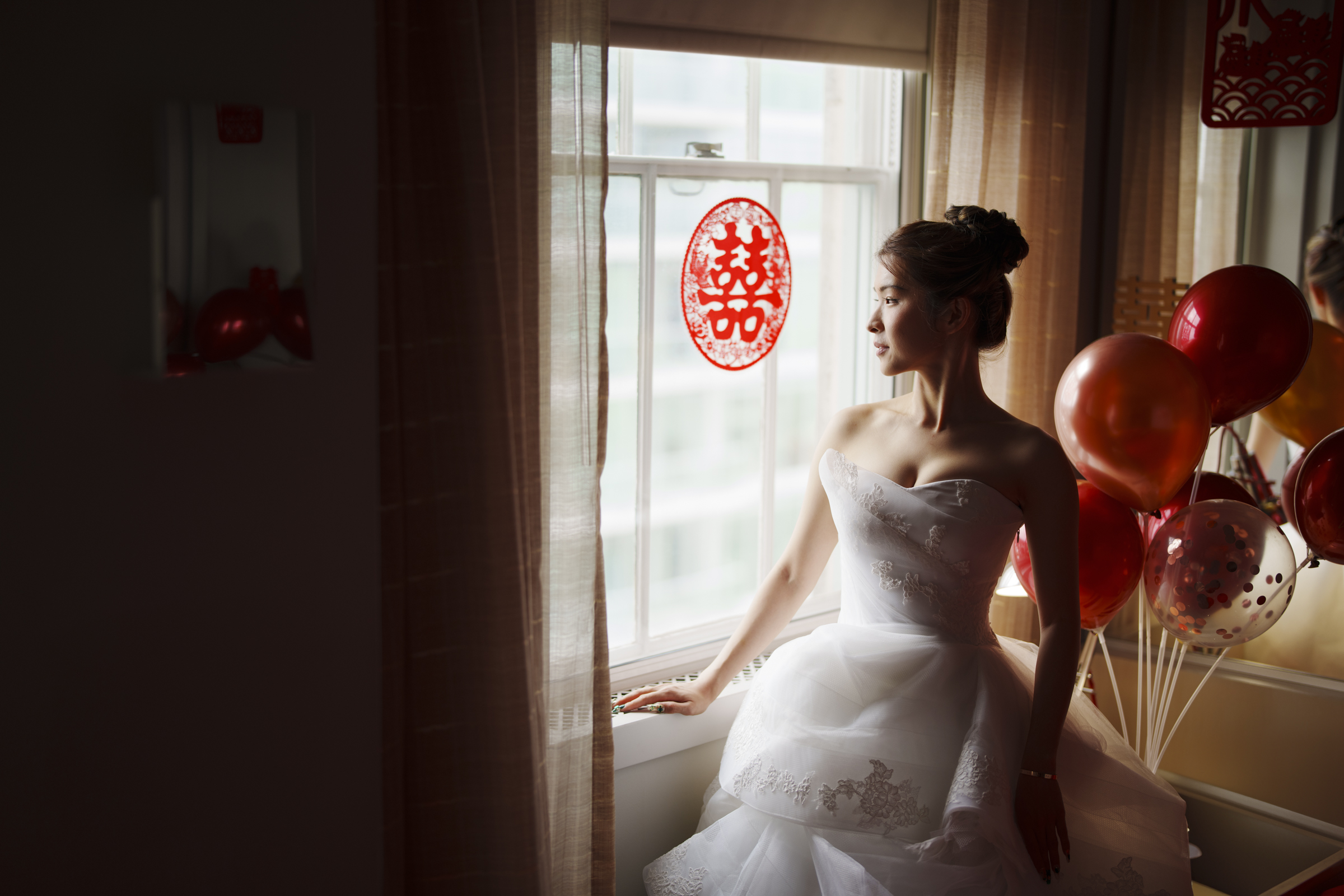 A bride in a strapless white gown stands near a window adorned with Chinese decorations, clutching a bouquet of red and orange balloons. Bathed in warm light, she gazes outside, dreaming of her Duke Chapel wedding.