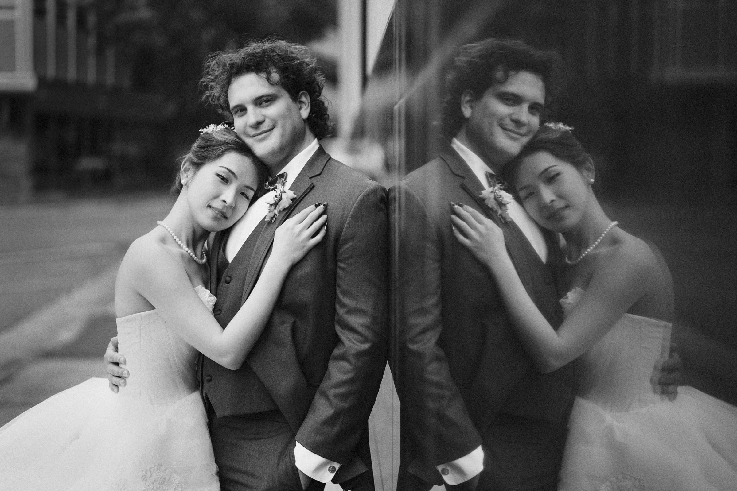 A black-and-white photo captures a Duke Chapel wedding moment as the bride leans affectionately on the groom's shoulder, both smiling softly. They stand against a reflective surface, creating a mirrored image of the couple.