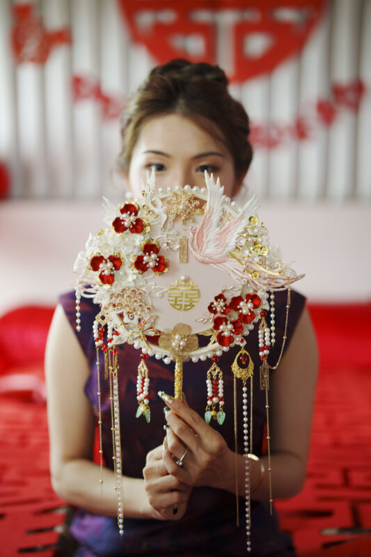 A woman holds an ornate fan decorated with flowers, beads, and a bird, partially covering her face. The backdrop resembles a Duke Chapel wedding with its rich red and white elements intertwined with Chinese symbols.