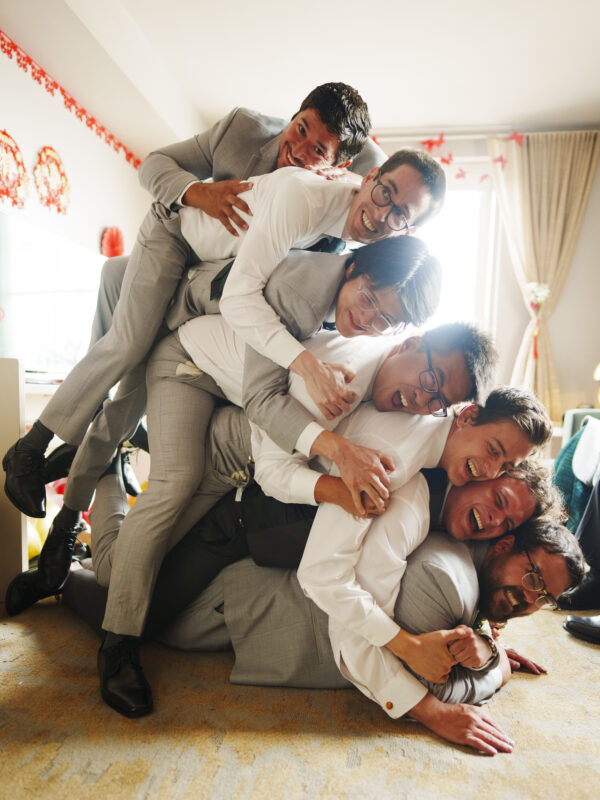 A group of men in formal attire are playfully piled on top of one another inside a brightly lit room with festive decorations, perhaps fresh from a Duke Chapel wedding. They appear to be laughing and enjoying the moment.