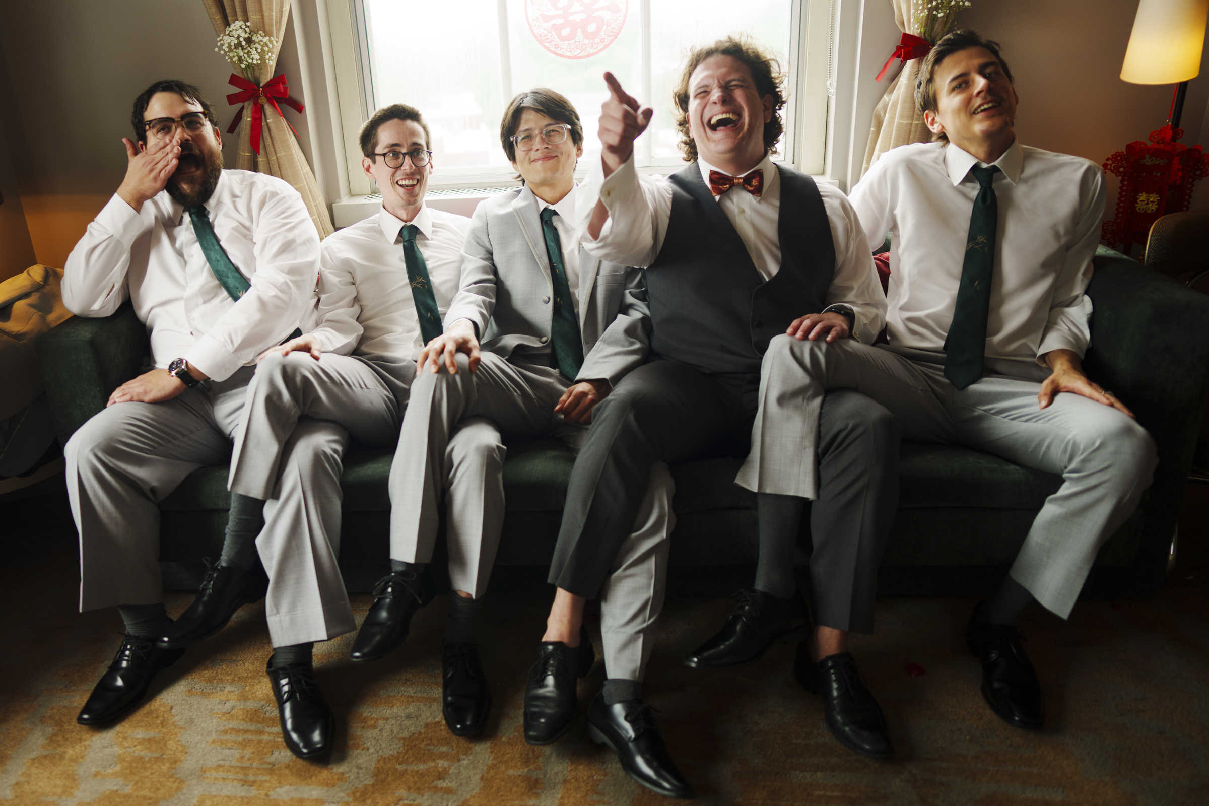 Five men in formal attire sit on a green couch, smiling and laughing, perhaps reminiscing about a Duke Chapel wedding. Four wear white shirts with green ties, while one dons a vest with a bow tie. They seem to be enjoying a light-hearted moment indoors.