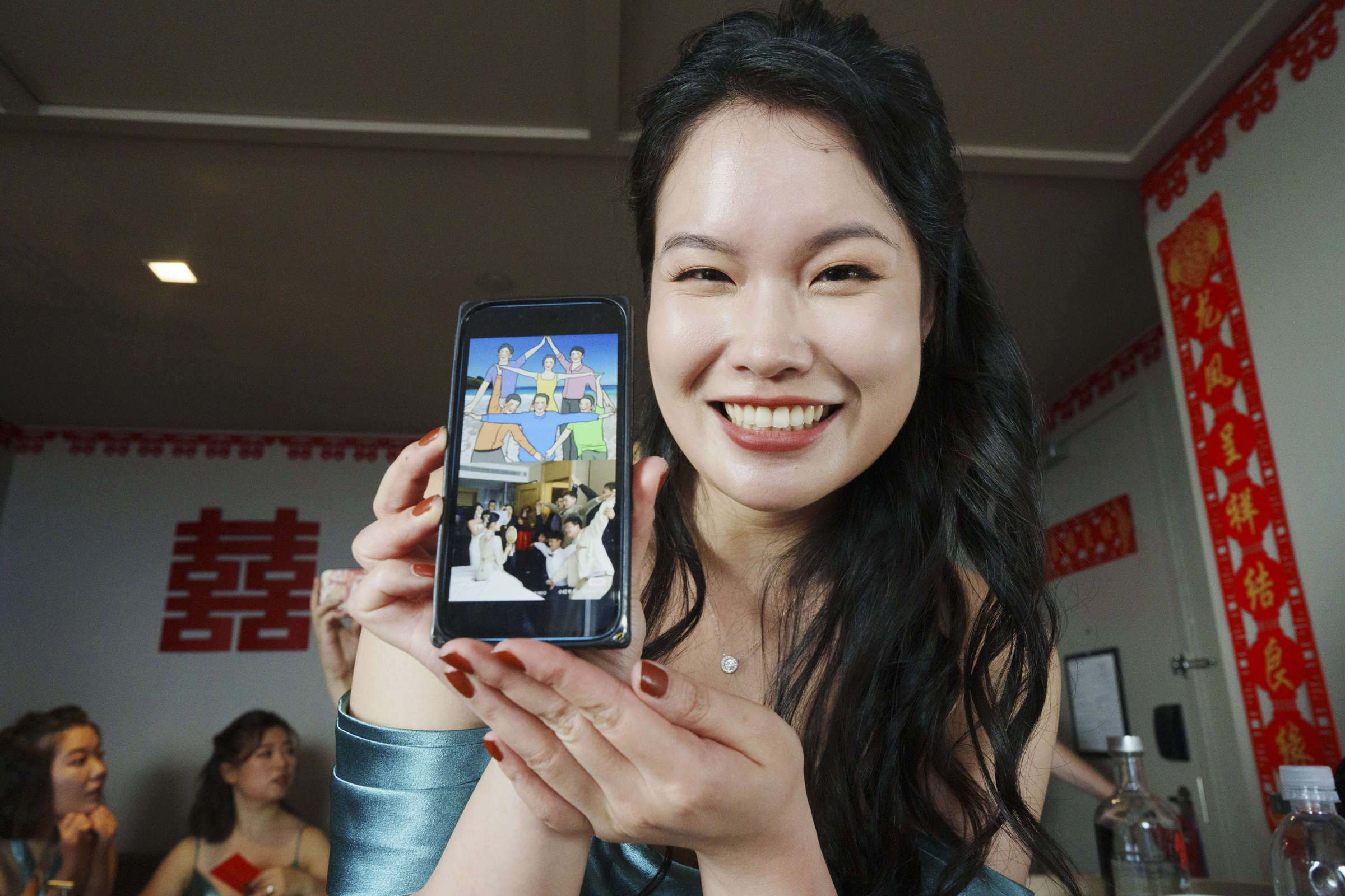 A smiling woman holds up a smartphone displaying a photo at a Duke Chapel wedding celebration. The room is adorned with red decorations, including a double happiness symbol. Several people are in the background, one laughing, in this festive setting.