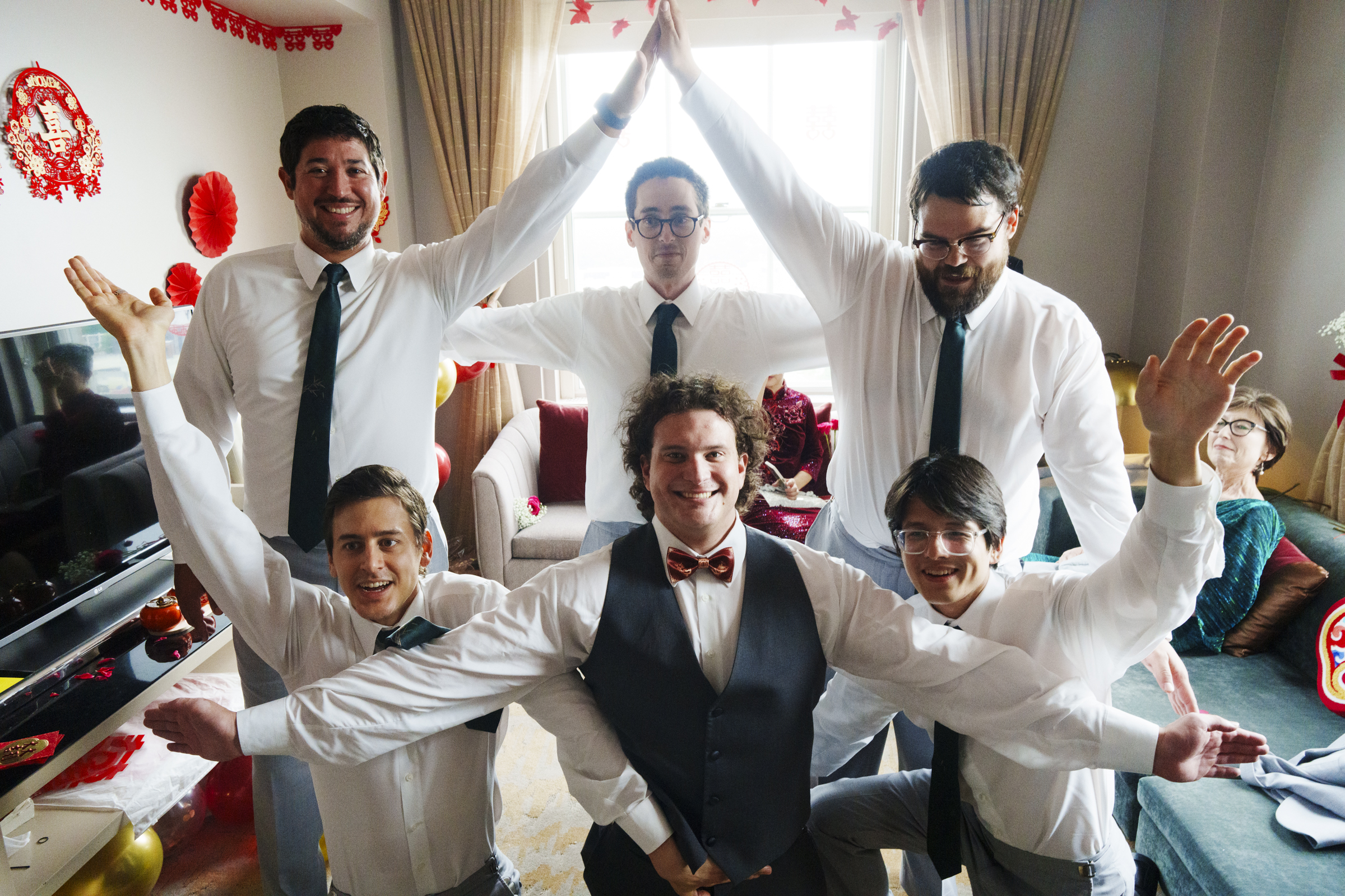 In a living room adorned with red decorations, a group of six, dressed in white shirts and ties—except one who sports a vest with a bow tie—form a pyramid shape. Their formation captures the spirit usually reserved for grand occasions like a Duke Chapel wedding, as someone smiles from the couch.