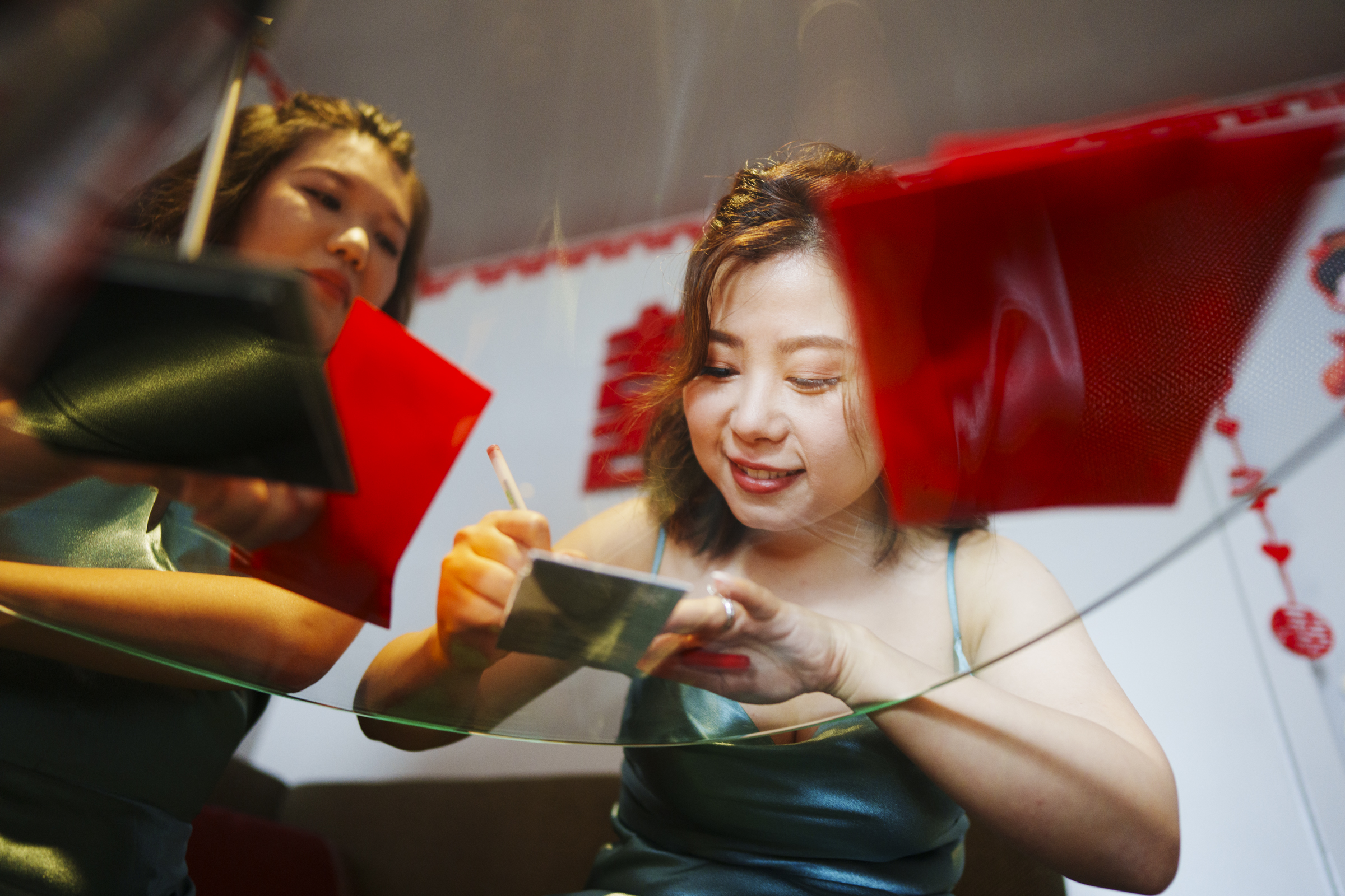 A woman smiles while jotting down notes at a table, surrounded by vibrant red decorations reminiscent of a Duke Chapel wedding. Her blue-green top complements the scene beautifully, and her reflection is captured in a nearby mirror.