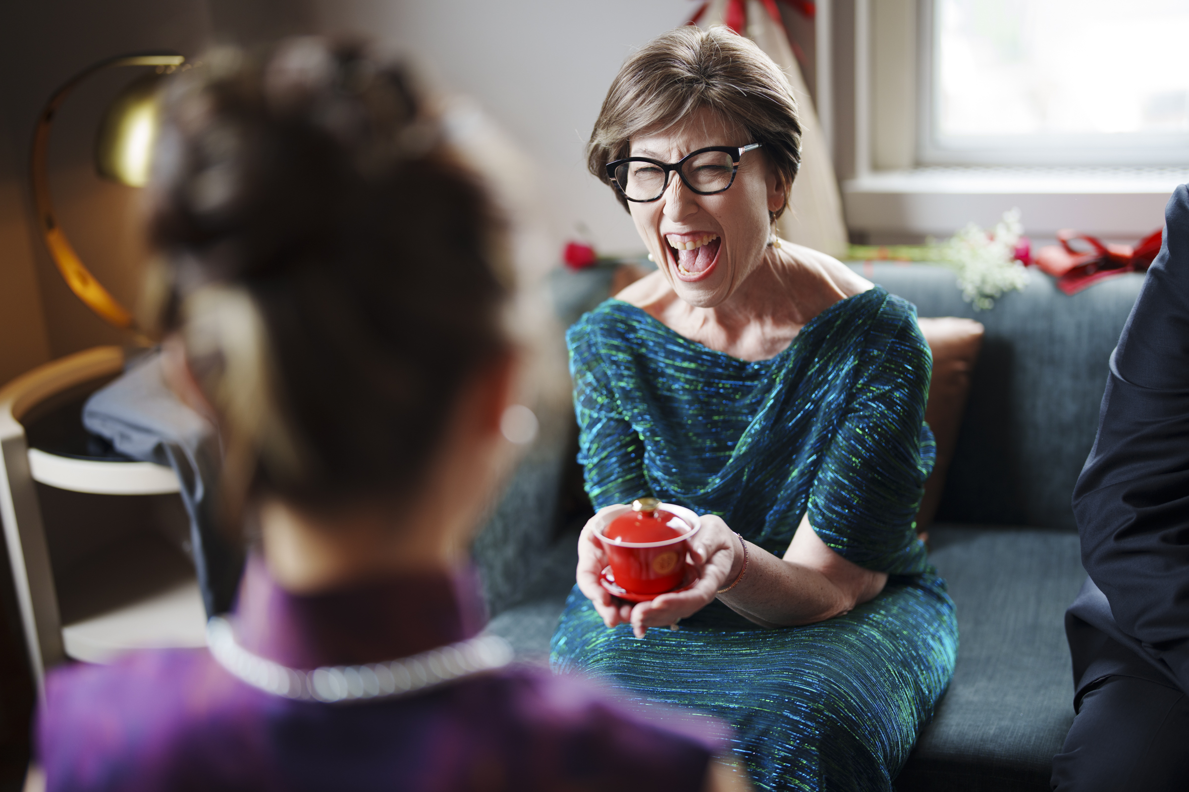 An older woman with glasses, in a blue dress, sits on a sofa laughing and holding a red cup. Her laughter fills the cozy room, evoking joy reminiscent of a Duke Chapel wedding. She faces another person whose back is to the camera, with light softly streaming through the window.