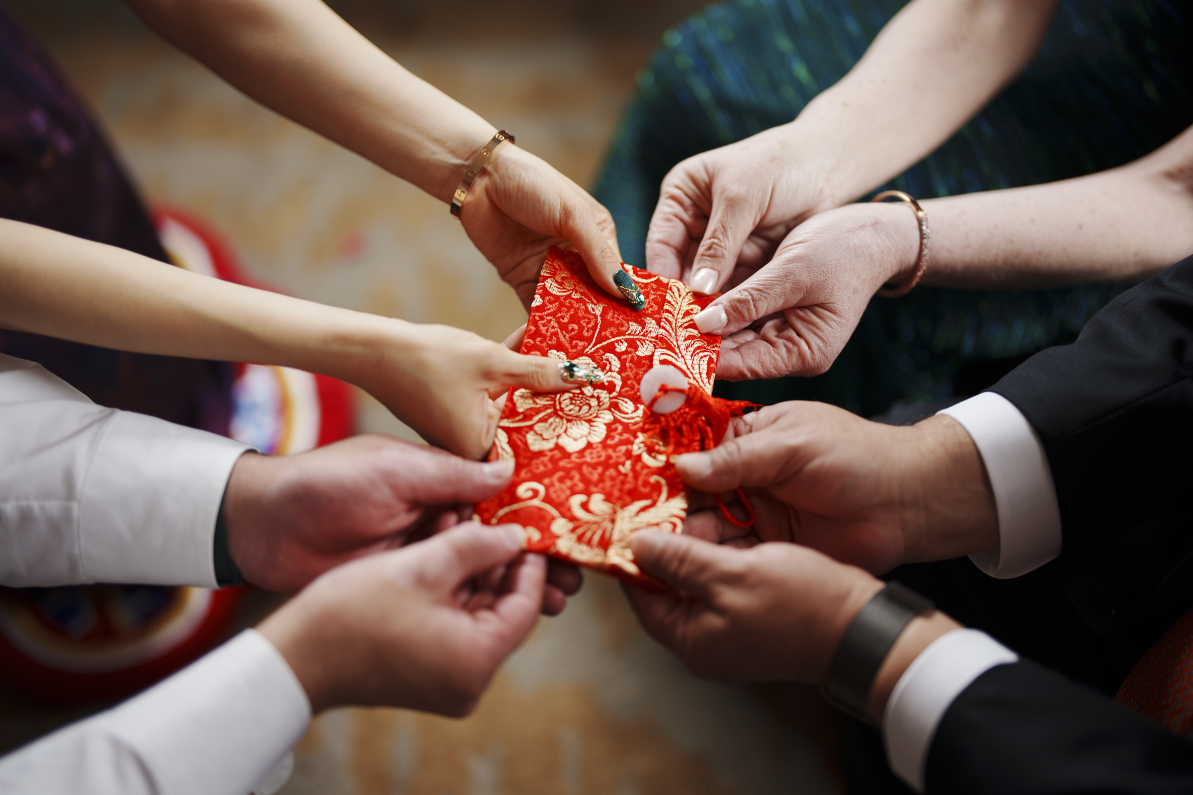 A group of people hold a red envelope with intricate gold patterns. The hands, belonging to various individuals, showcase diversity in skin tones. This scene suggests a celebratory exchange, possibly reminiscent of the vibrant traditions at events like a Duke Chapel wedding.