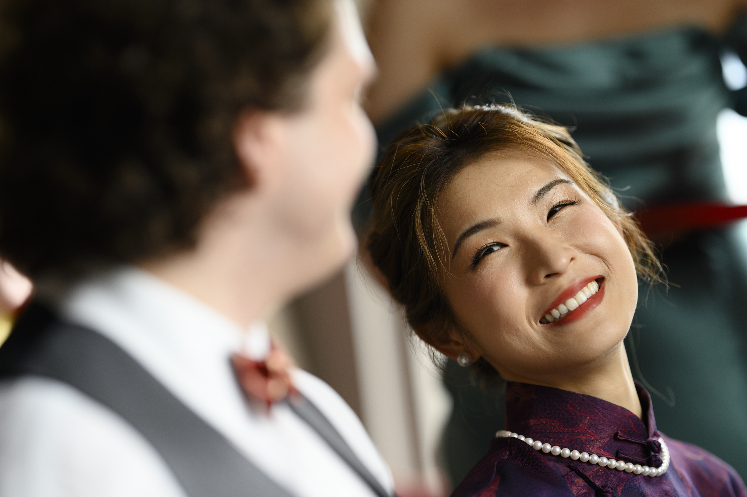 A woman in a purple dress with a pearl necklace smiles warmly at a person in a white shirt, vest, and red bow tie. Slightly out of focus, they are likely enjoying the joyous ambiance of a Duke Chapel wedding celebration.