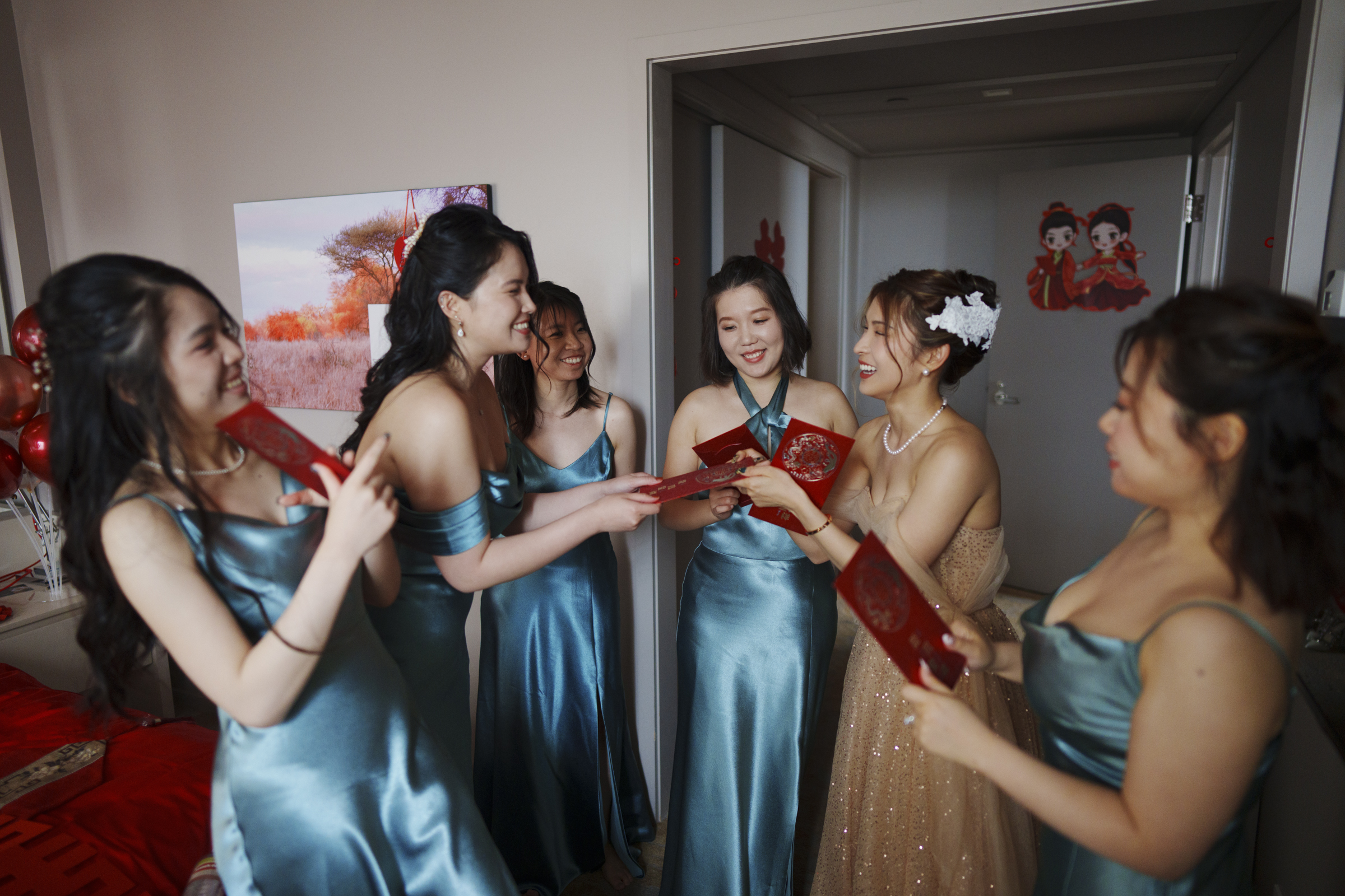 At a Duke Chapel wedding, a bride in a gold dress joyfully distributes red envelopes to five women in matching teal dresses. They gather in a festively decorated room, sharing a light-hearted moment together, creating memories that will last a lifetime.