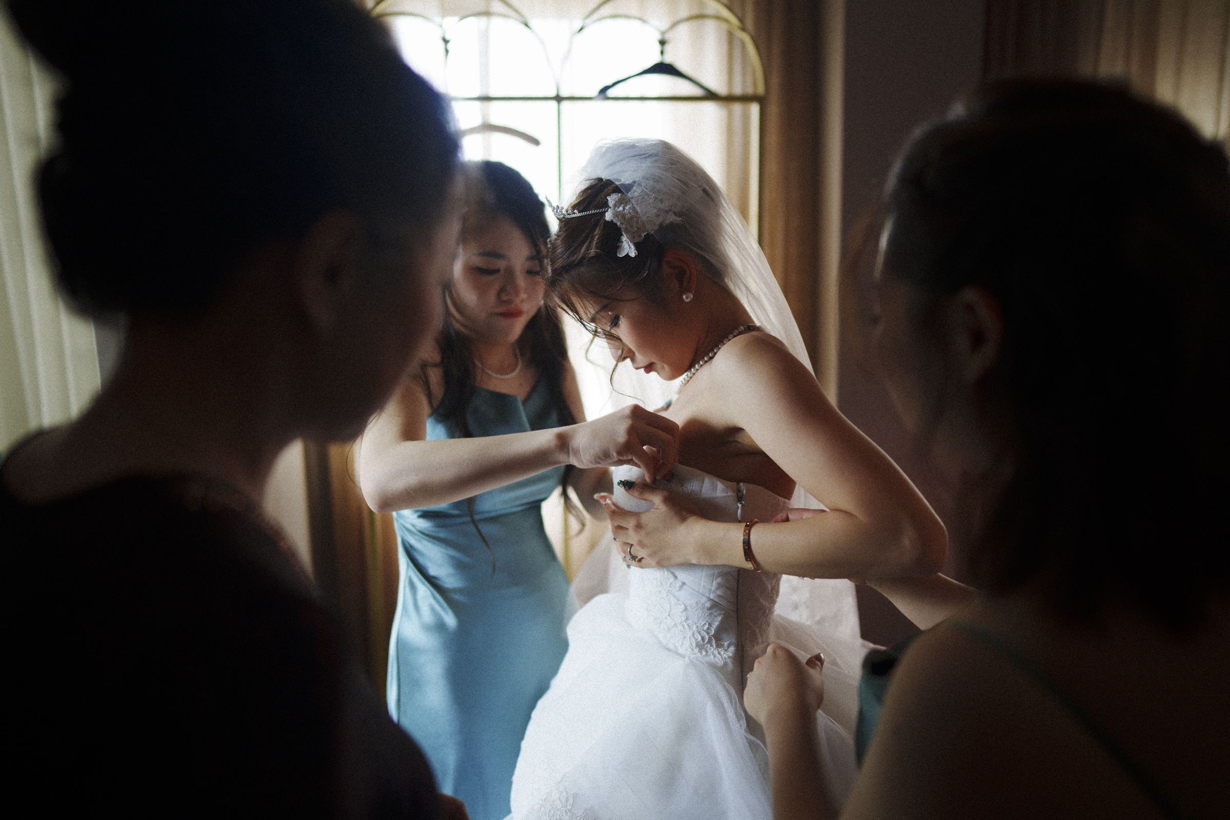 In a softly lit room, the bride in her white gown is lovingly assisted by three women as she prepares for her Duke Chapel wedding. With a window in the background, the atmosphere remains intimate and focused.