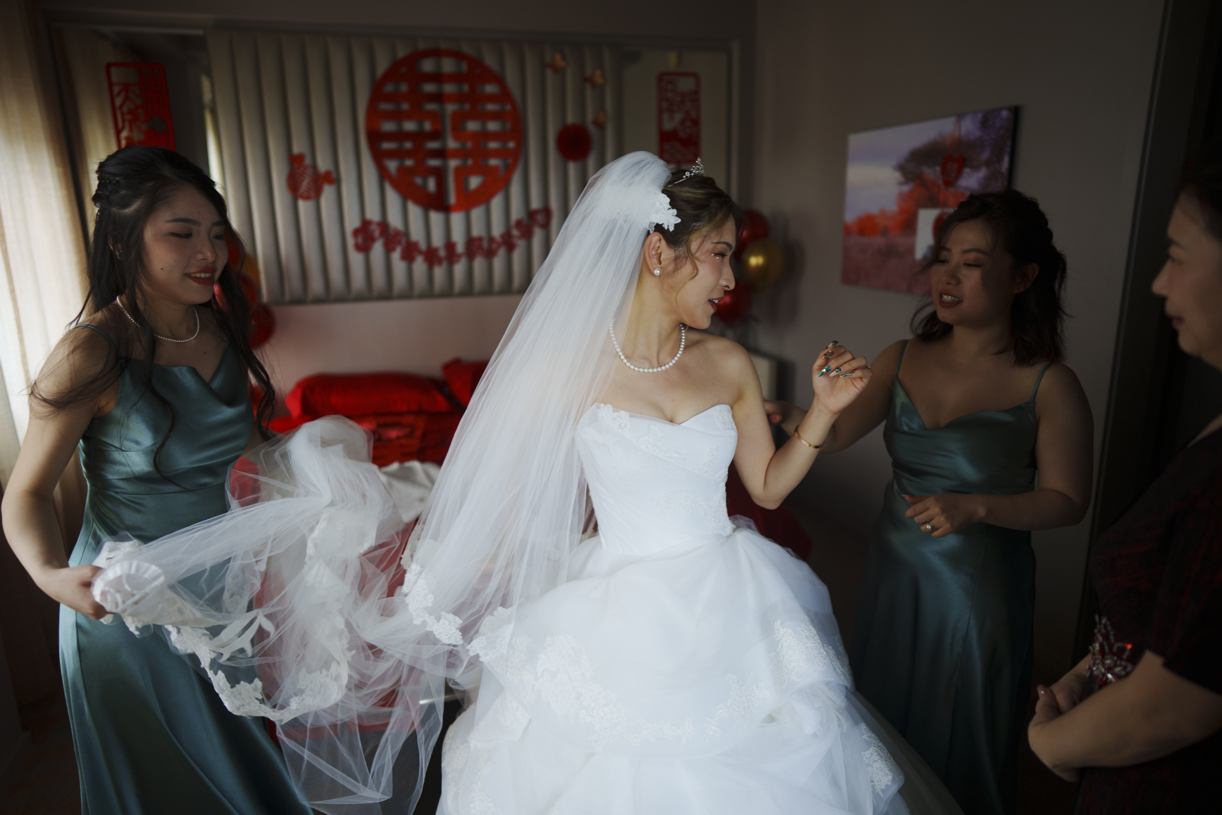 A bride in a white wedding dress and veil stands with three bridesmaids in green dresses. They are gathered in a dimly lit room adorned with red symbols, preparing for the Duke Chapel wedding ceremony.