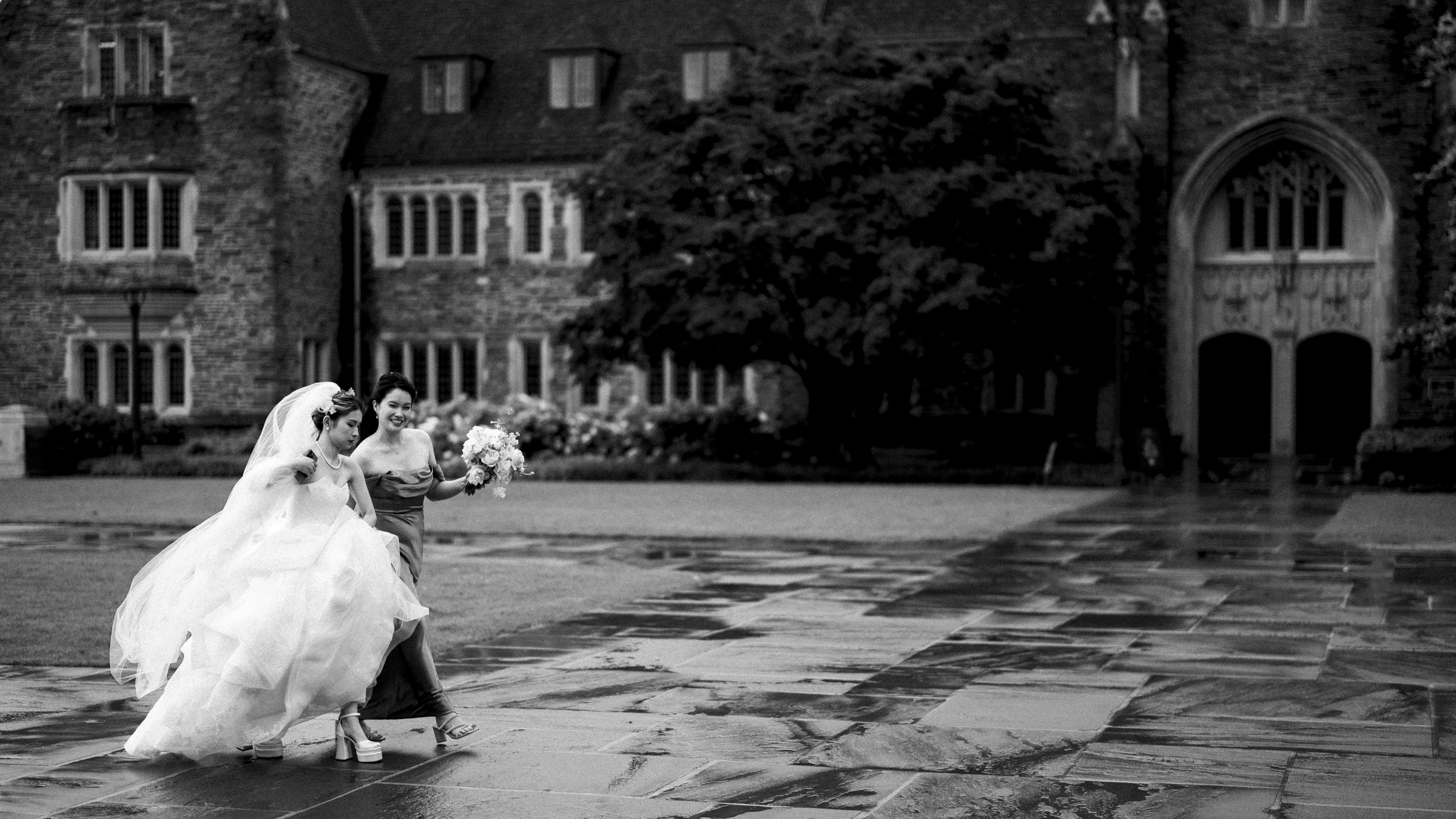 A black and white photo captures a Duke Chapel wedding scene: a bride lifting her dress, accompanied by her bridesmaid holding a bouquet. They stroll through the rain in front of the chapel's majestic stone facade, with its arched windows and grand entrance adding timeless elegance to the moment.