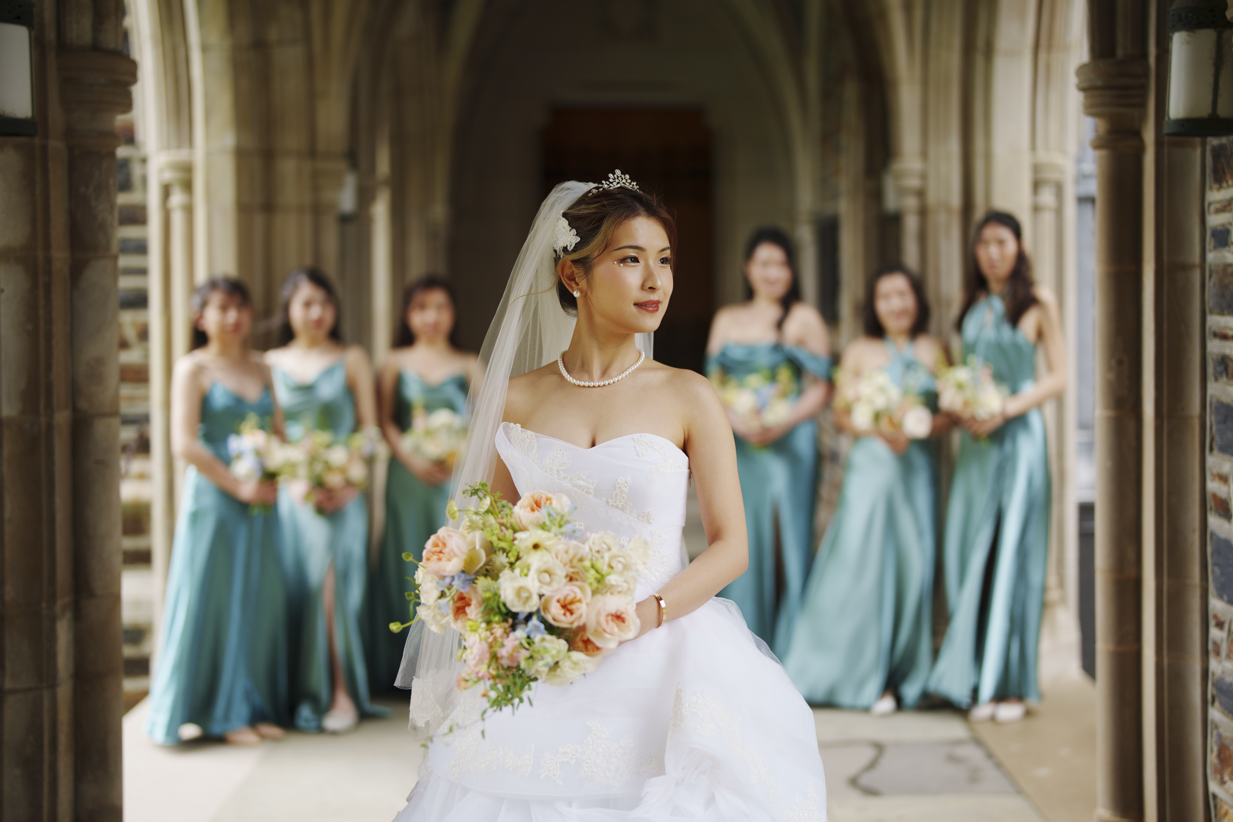 A bride in a white gown and veil holds a bouquet of flowers at a Duke Chapel wedding, standing gracefully in front of five bridesmaids dressed in teal gowns. They pose in the elegant, arched corridor, creating a joyful and picturesque scene.