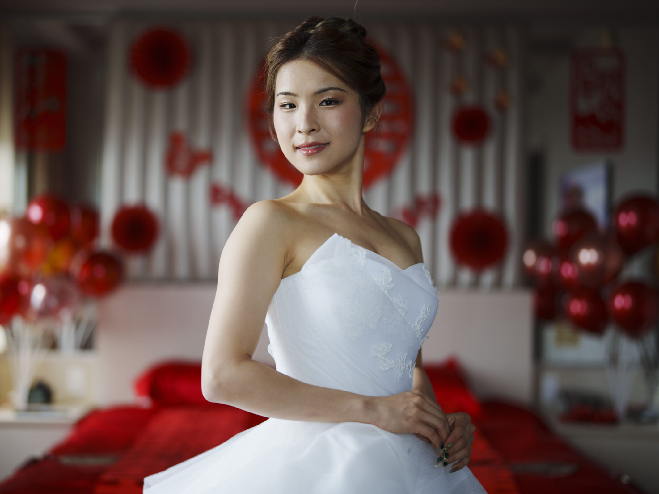 A bride in a white wedding dress stands in a room adorned with red and white balloons, capturing the elegance of a Duke Chapel wedding. She smiles slightly and poses gracefully, her hair styled up, while the background is softly blurred.