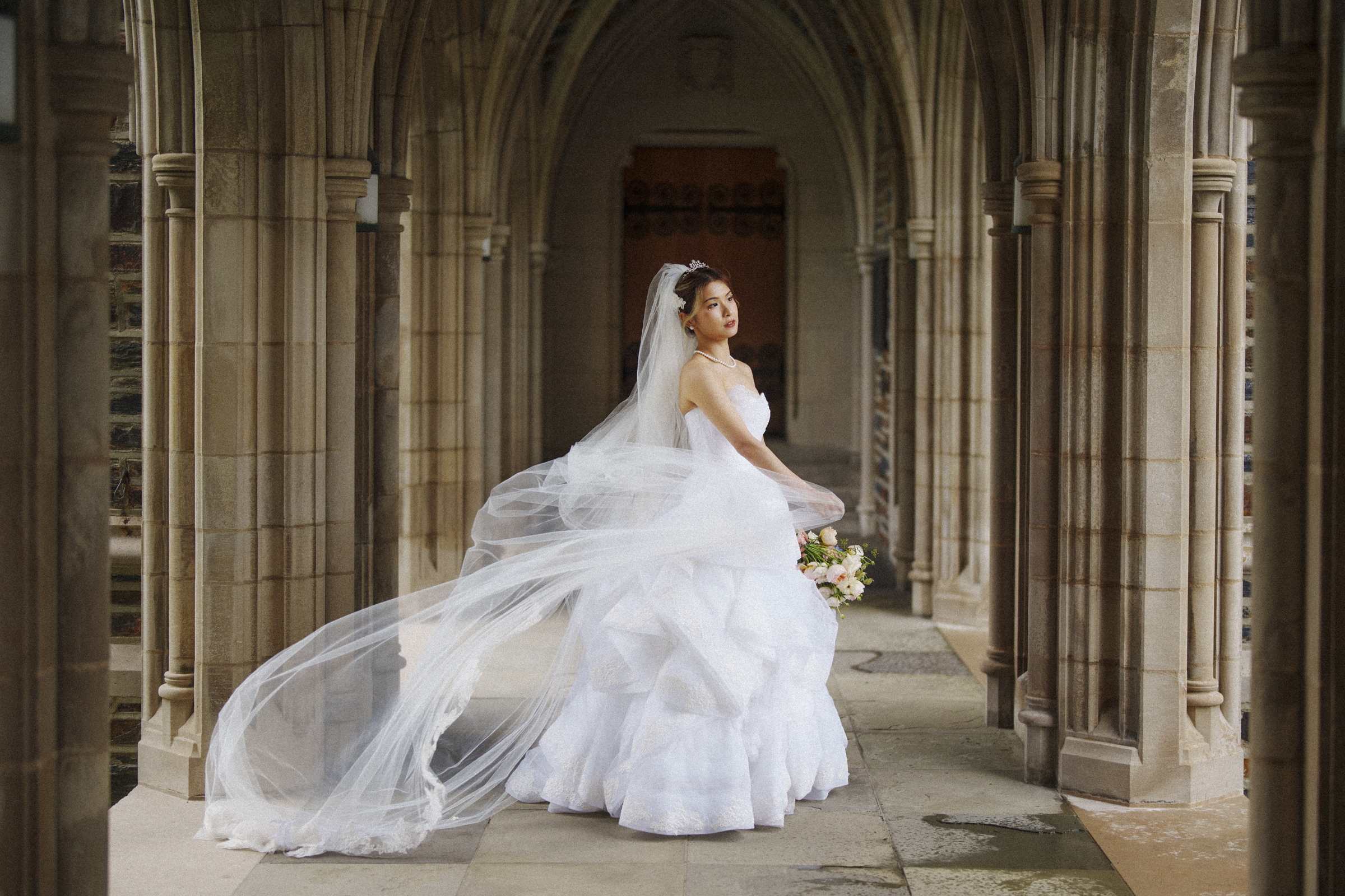 The bride in a white gown and long veil stands gracefully in the stone archway of Duke Chapel, holding a bouquet and glancing over her shoulder. The serene scene is elegant, with soft lighting accentuating the architectural details.