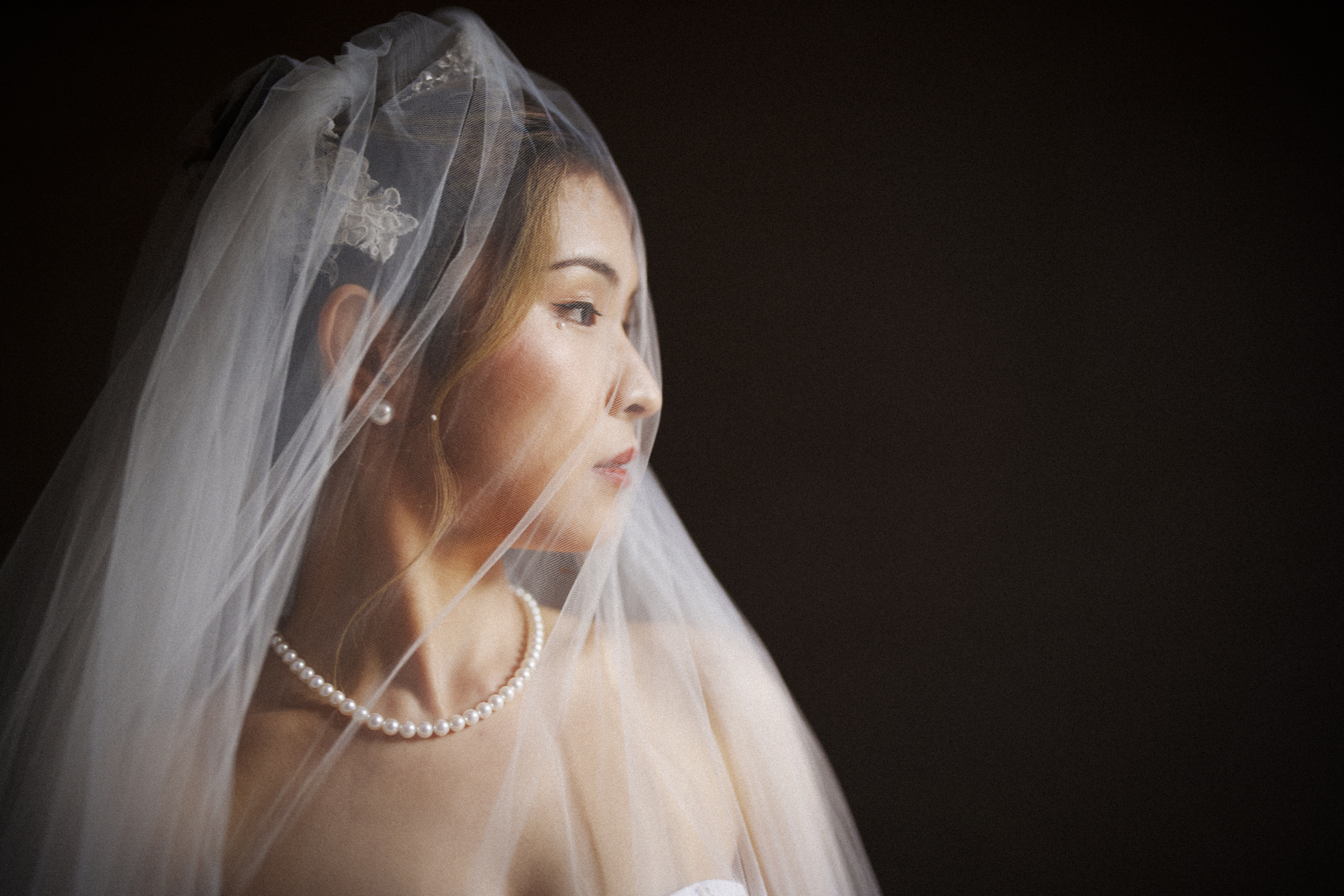 A bride in a white veil and pearl necklace gazes to the side, embodying the elegance of a Duke Chapel wedding. Her hair is styled gracefully, and her subtle makeup complements the dark background, highlighting her serene and contemplative expression.