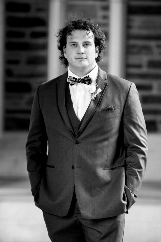 A person wearing a suit, vest, and bowtie stands confidently outdoors near Duke Chapel. The background is blurred, highlighting the individual’s formal attire and curly hair. The black and white photo gives it a classic wedding feel.