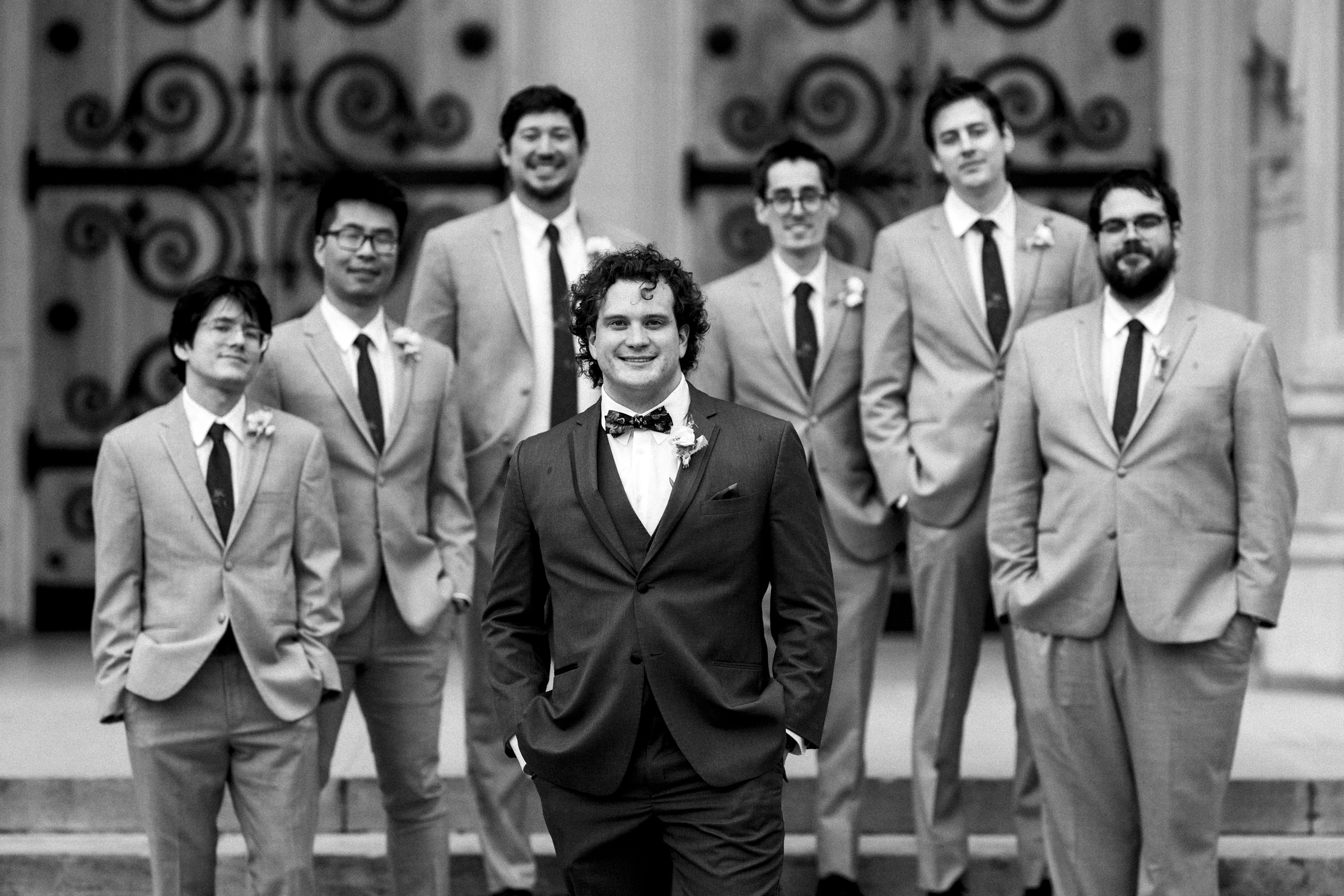 Seven men in suits, one standing in the foreground with a bow tie, smiling. The group is positioned on steps with ornate doors reminiscent of a Duke Chapel wedding in the background. Black and white photo.