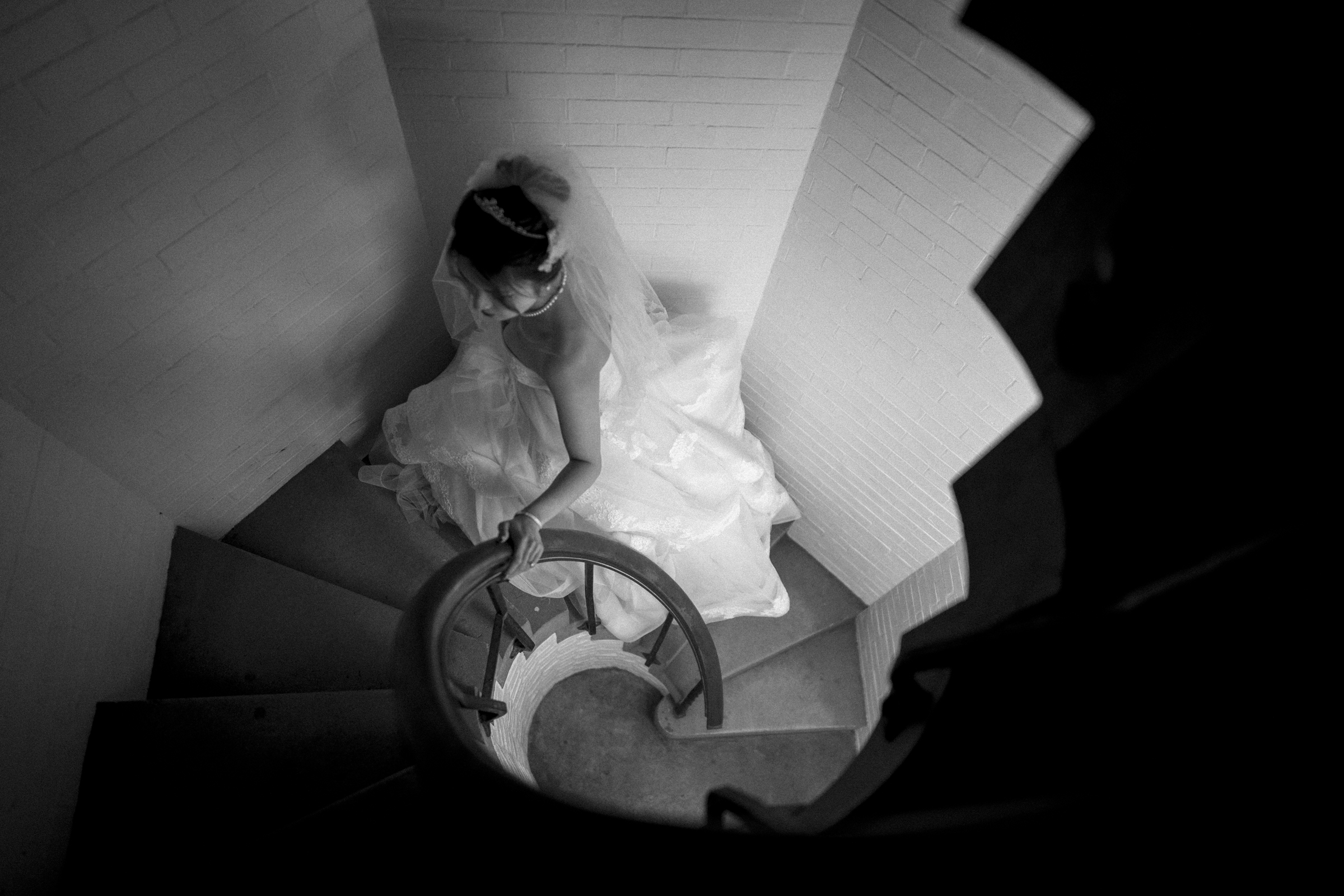 A bride in a white gown and veil gracefully descends a spiral staircase, holding the handrail. This black and white photo, taken at a Duke Chapel wedding, captures the elegant swirl of her dress against the brick walls, creating a romantic and timeless atmosphere.