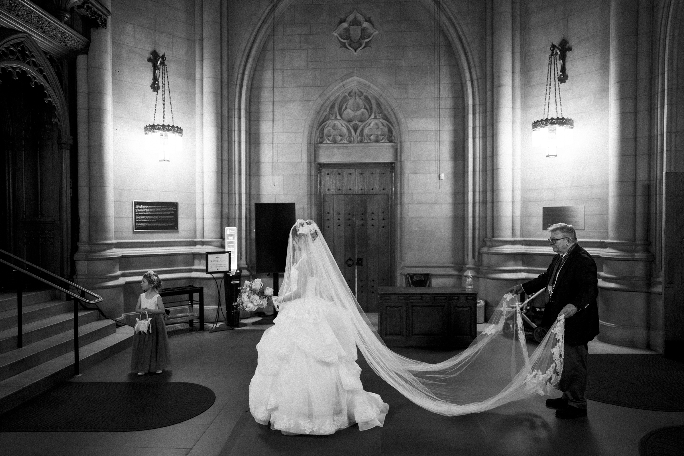 A bride in a voluminous wedding gown and veil stands in the grand interior of Duke Chapel. A man assists with her long veil, while a young girl in a dress looks on. The setting is elegant, with tall stone walls and ornate details. Black and white photo capturing timeless grace.