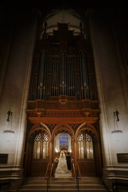 A bride and groom stand at the altar inside the grand cathedral of Duke Chapel, facing an ornate wooden structure with large organ pipes. The dim lighting elegantly highlights the architecture and stained glass window above them, creating a stunning backdrop for their wedding ceremony.