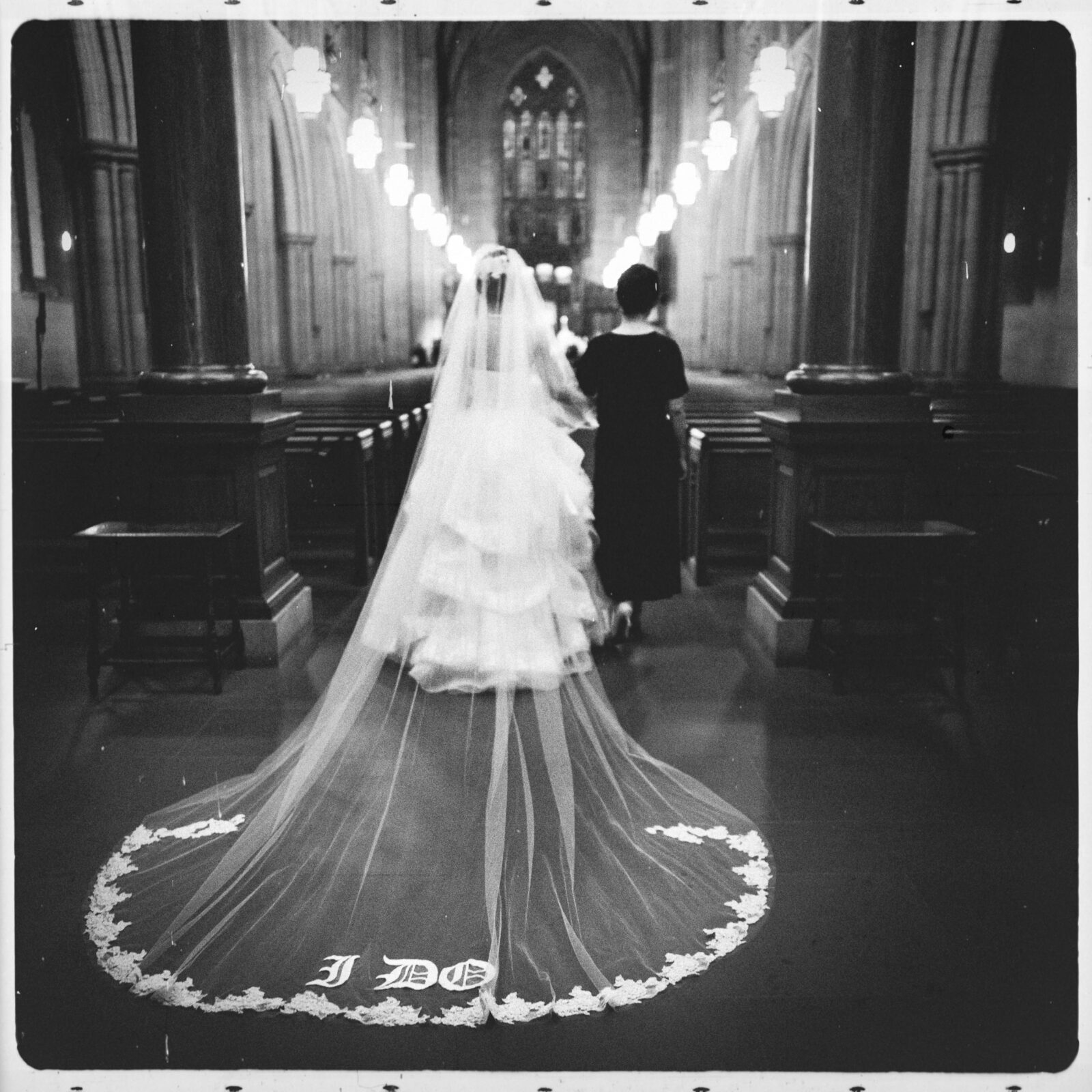 The bride and woman walk down the aisle of Duke Chapel. Her long veil, elegantly spread on the floor, is embroidered with the words "I DO" in white. The dimly lit cathedral interior highlights ornate columns and arched ceilings, adding a touch of grandeur to this memorable moment.