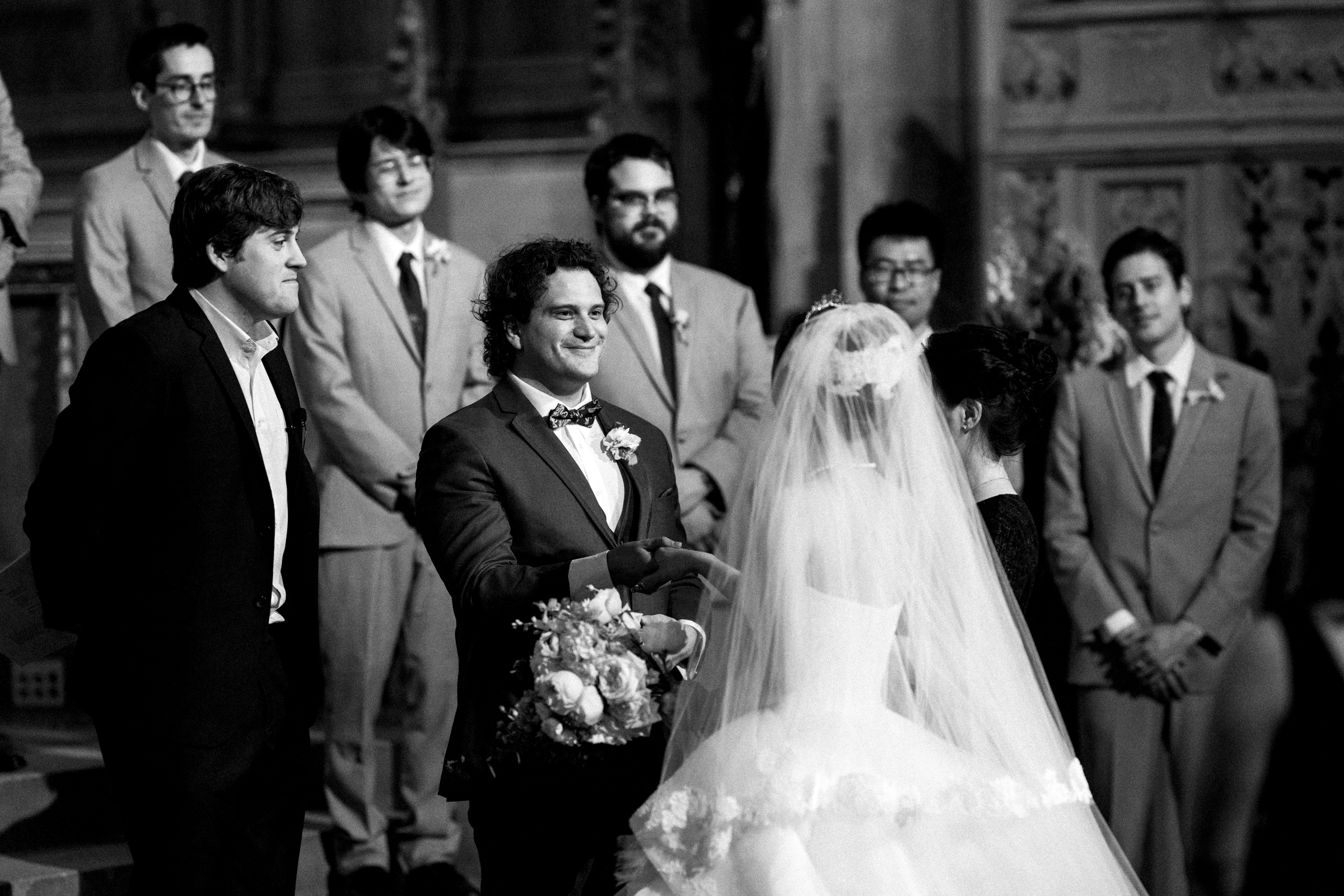 A black-and-white photo captures a Duke Chapel wedding ceremony. The groom, holding a bouquet, smiles at the bride beneath her veil. Several groomsmen stand behind them in their suits, bearing witness to this timeless moment.