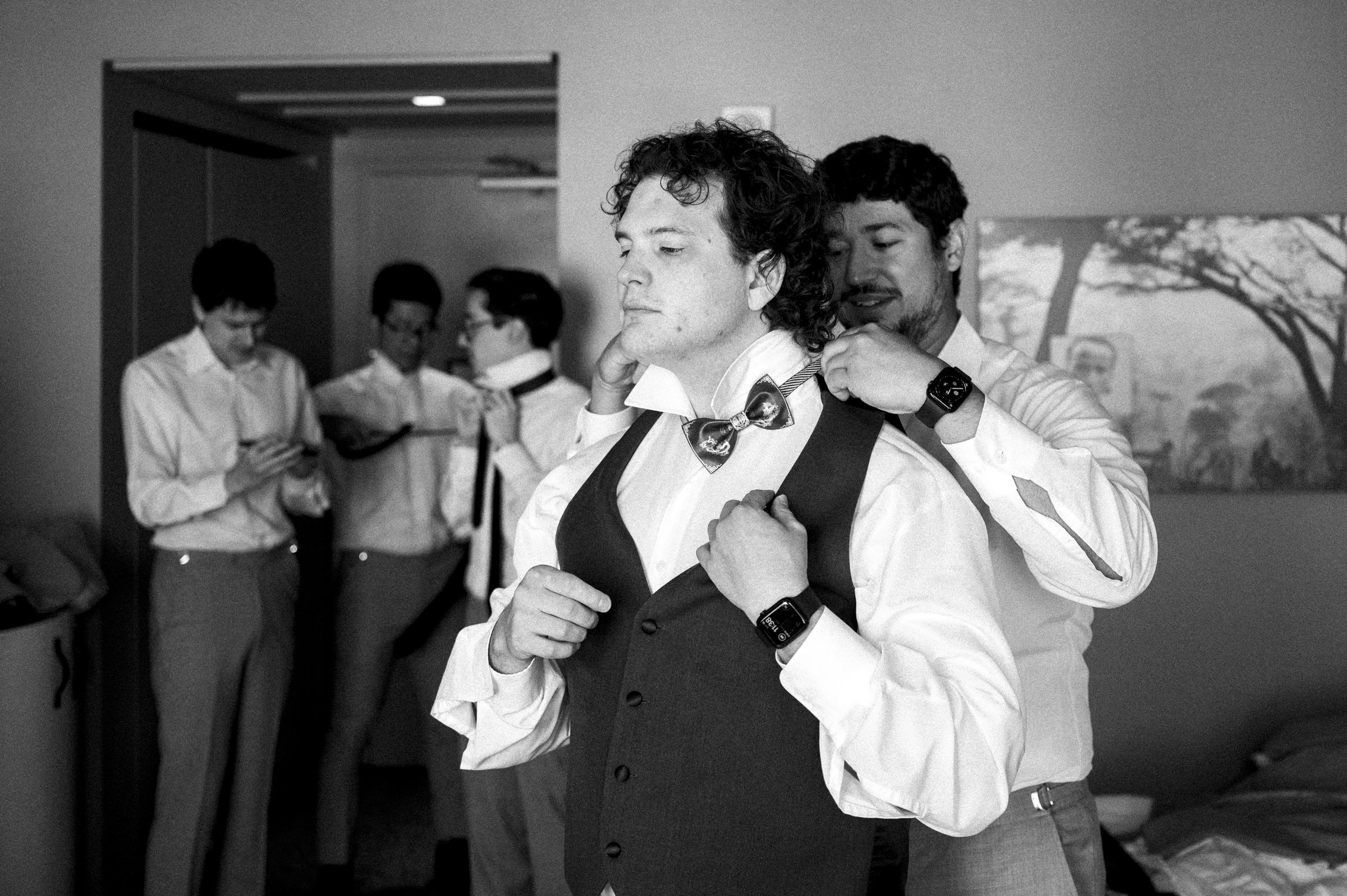 A groom, preparing for his Duke Chapel wedding, adjusts his vest while a groomsman assists with the bow tie. In the softly lit room adorned with a painting, four other groomsmen get ready in the background. The timeless elegance of this black and white photo captures the moment perfectly.