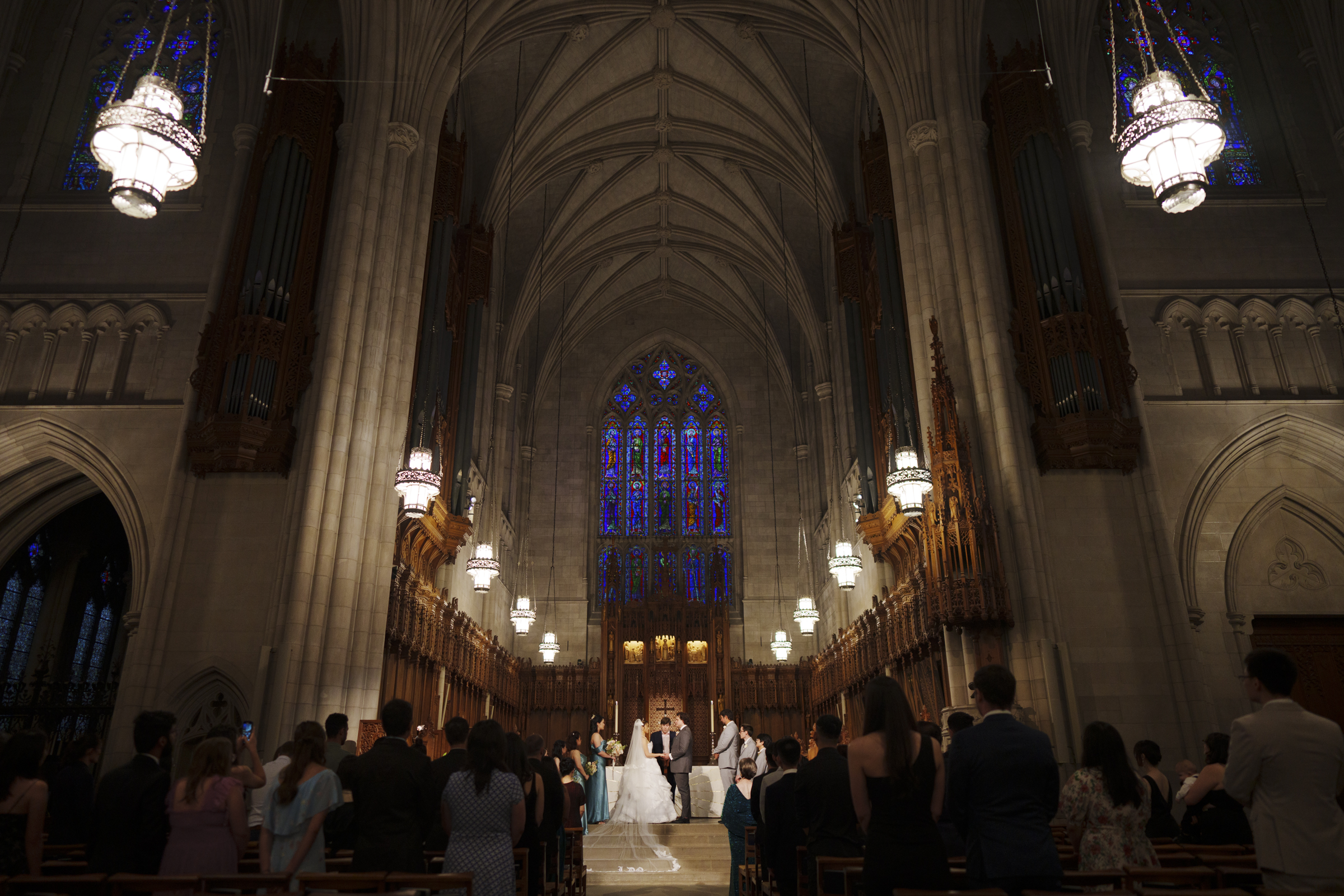 A Duke Chapel wedding unfolds in a grand cathedral with high vaulted ceilings, ornate chandeliers, and large stained glass windows. The bride and groom stand at the altar, surrounded by guests seated in pews.