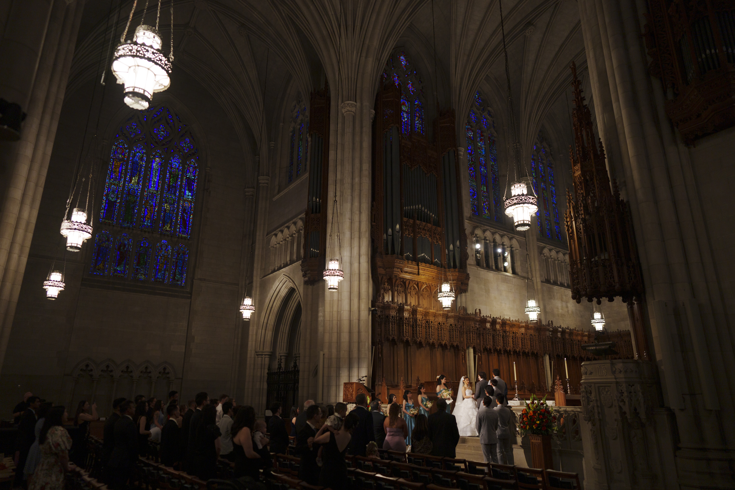 A Duke Chapel wedding unfolds in a grand church with high vaulted ceilings and stained glass windows. Guests are seated, and the couple stands at the altar under ornate lighting. The dim interior accentuates the architectural details, creating an unforgettable atmosphere.