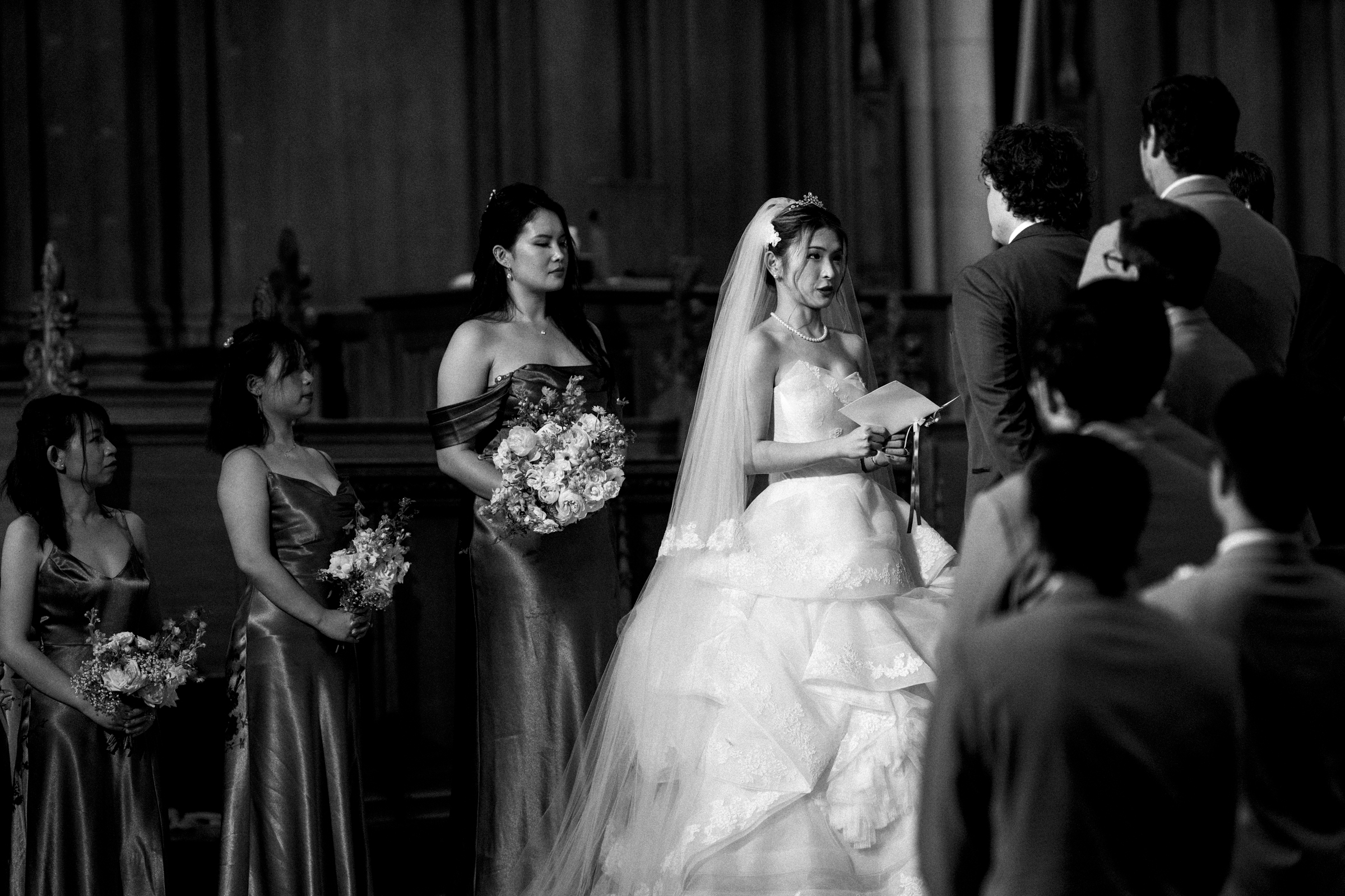 In a black and white photo of a Duke Chapel wedding, the bride, in a long gown and veil, stands holding a paper. Bridesmaids clutch bouquets as the groom in his suit faces her, with guests looking on in the church's grand setting.