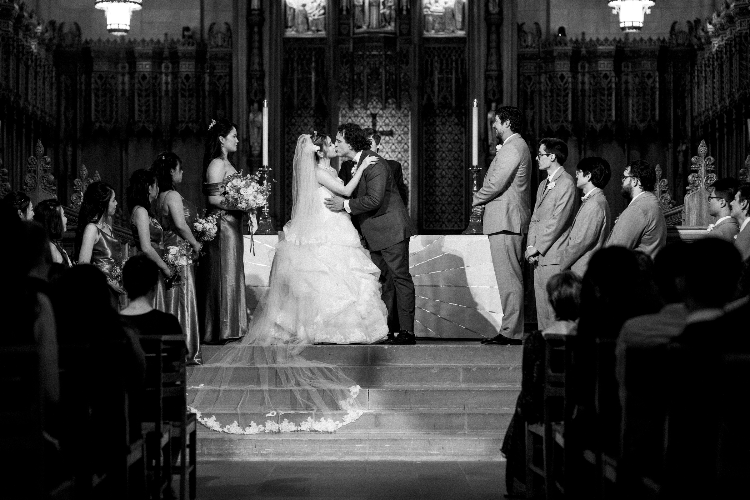 A black and white photo captures a Duke Chapel wedding ceremony. The bride and groom share a kiss at the altar, embraced by their bridal party. They stand before a beautifully decorated backdrop, with candles and floral arrangements lining the aisle. Guests are seated in pews, witnessing this precious moment.