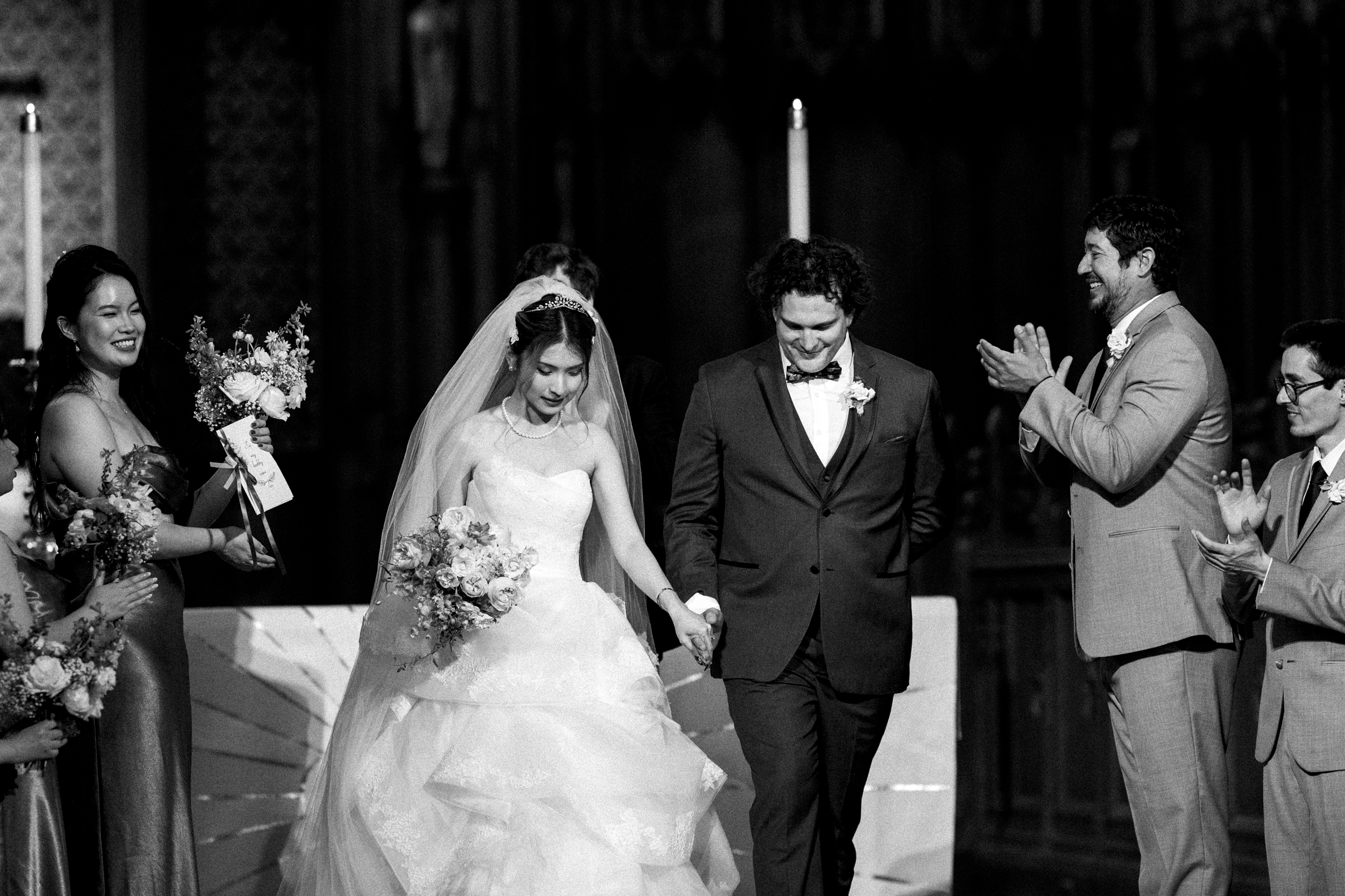 A black-and-white photo captures a Duke Chapel wedding ceremony, with the bride and groom smiling as they hold hands. The bride clutches a bouquet, surrounded by joyful guests, including bridesmaids and groomsmen, all clapping and celebrating the couple's happiness.