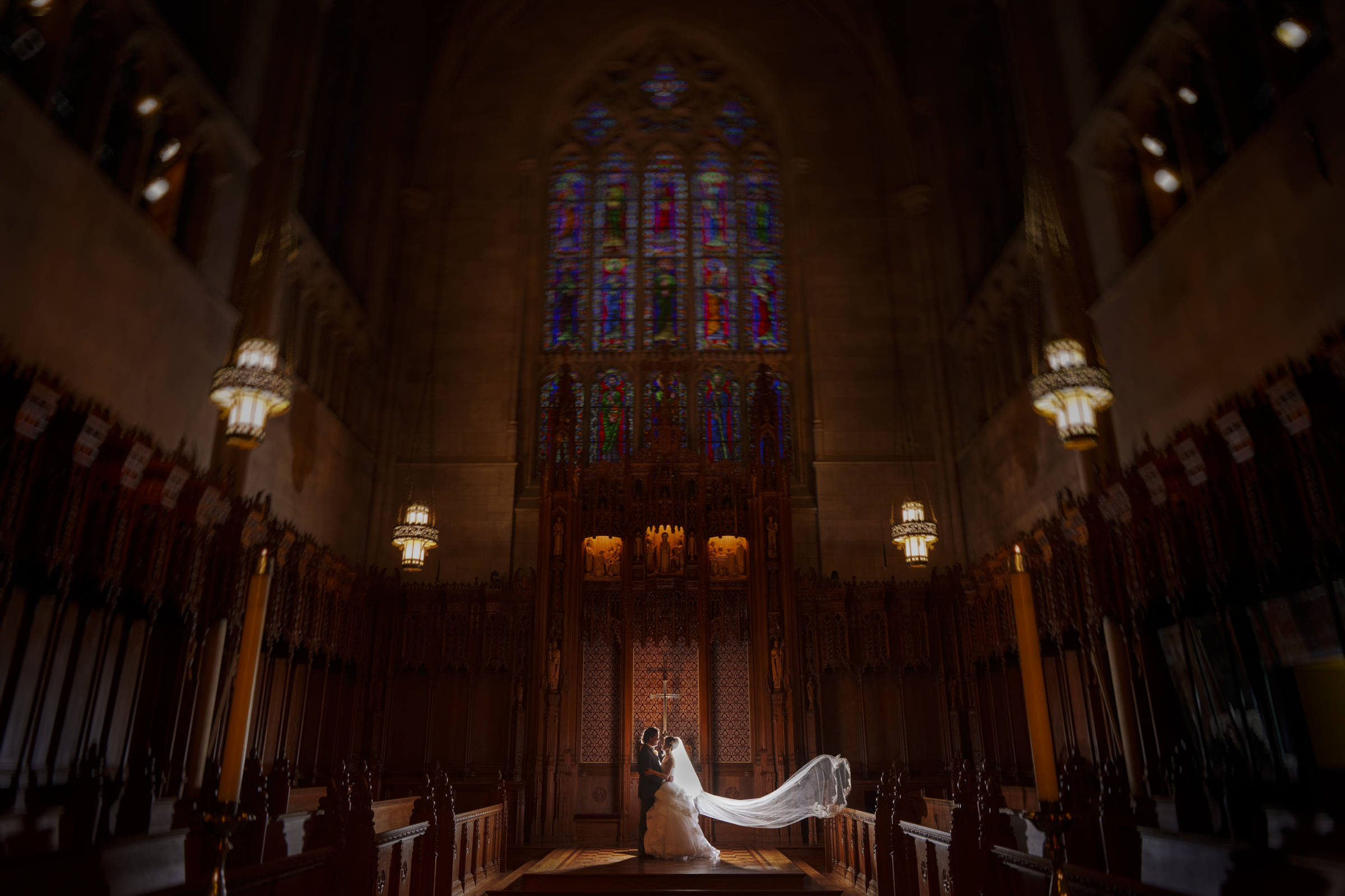 A bride and groom stand in the ornate, dimly lit Duke Chapel with a long, flowing veil trailing behind the bride. Stained glass windows and intricate woodwork enhance the grandeur of this wedding scene.