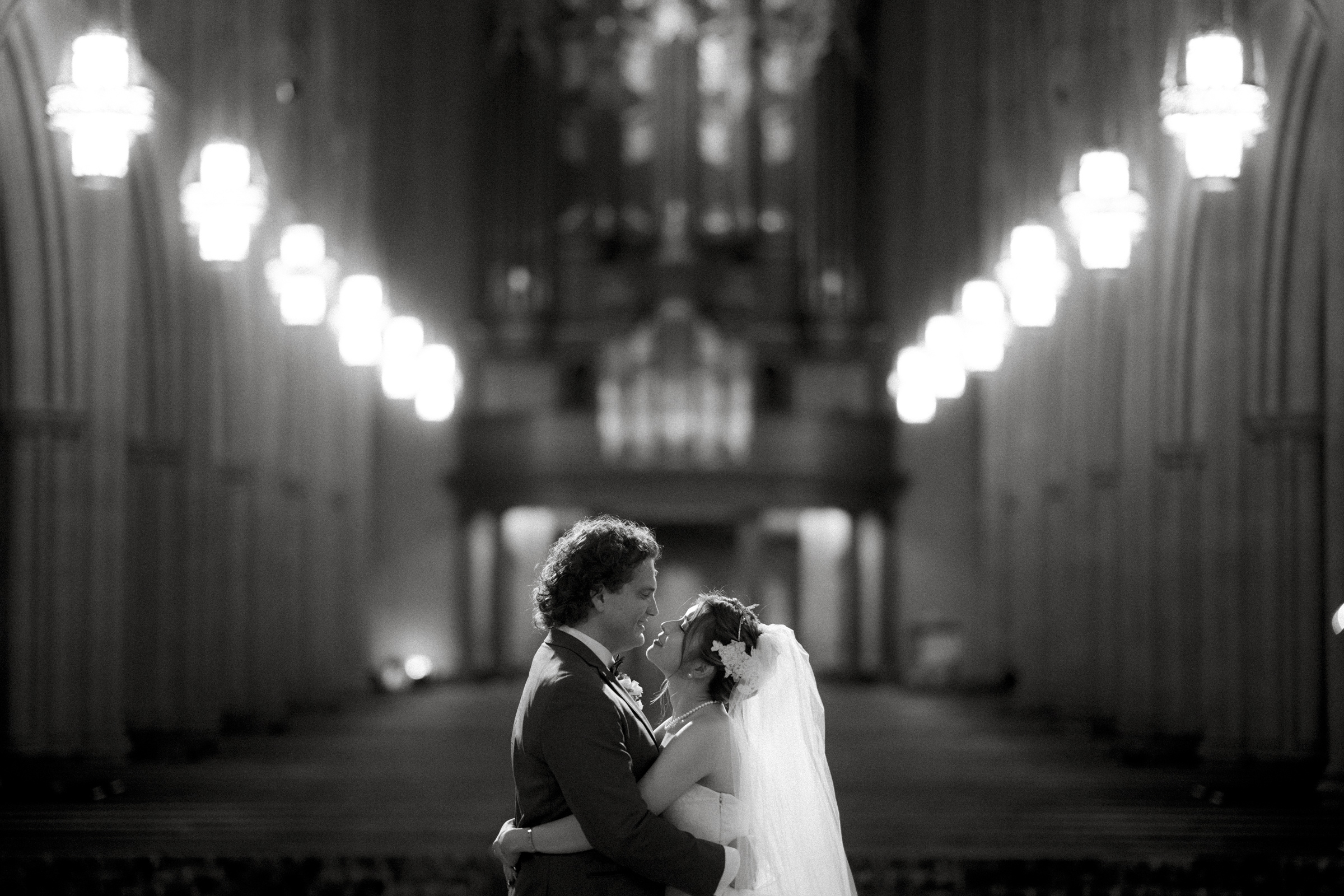 A couple in wedding attire embrace and smile at each other in the aisle of the grand Duke Chapel. Rows of glowing chandeliers illuminate the ornate, arched interior in this dimly lit, black and white photograph.