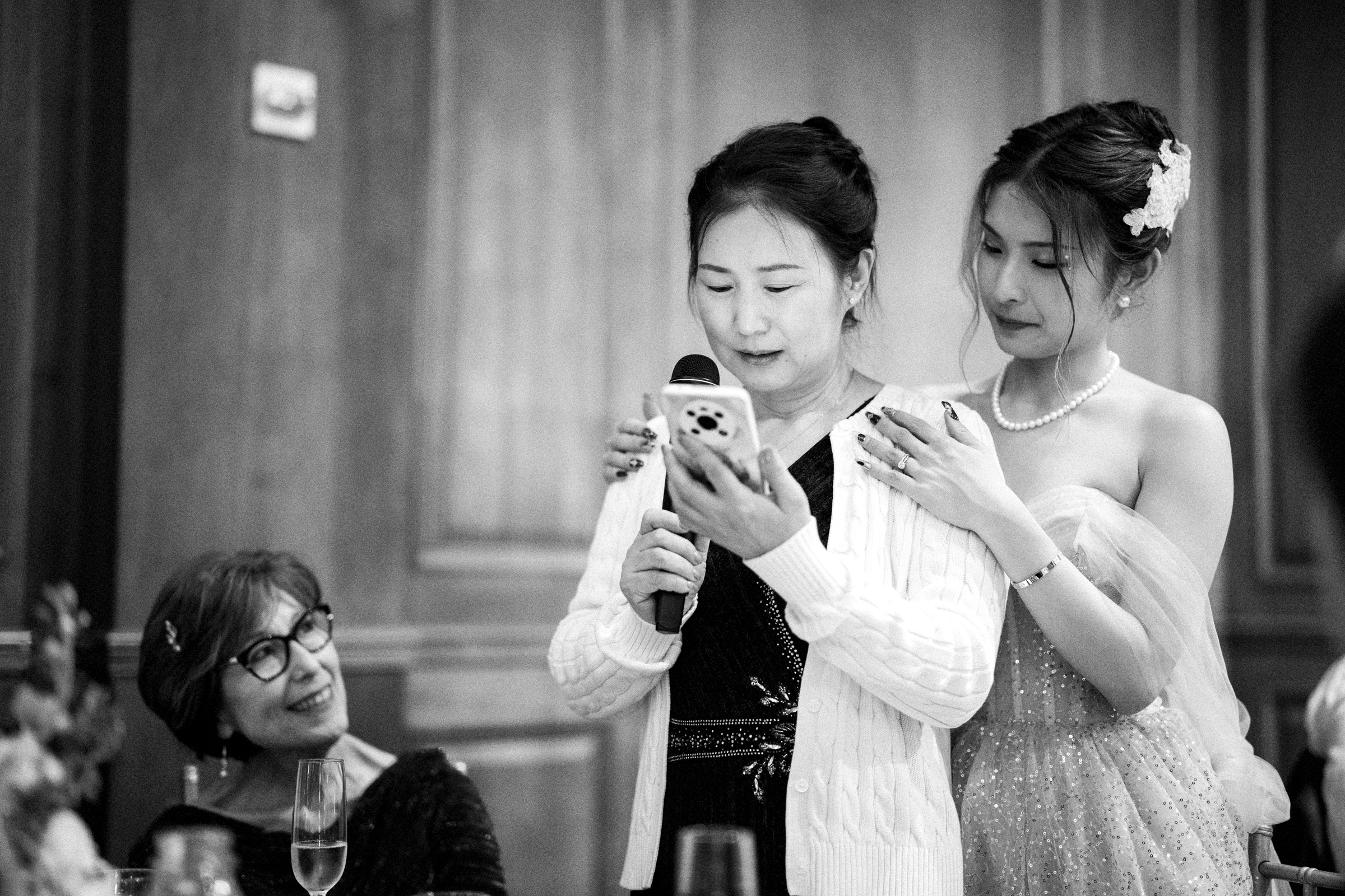 A woman in a white cardigan speaks into a microphone at a Duke Chapel wedding, reading from her phone as a younger woman in a strapless dress gently holds her shoulders. Another seated woman watches them, smiling. They are in a wood-paneled room adorned with floral decorations.