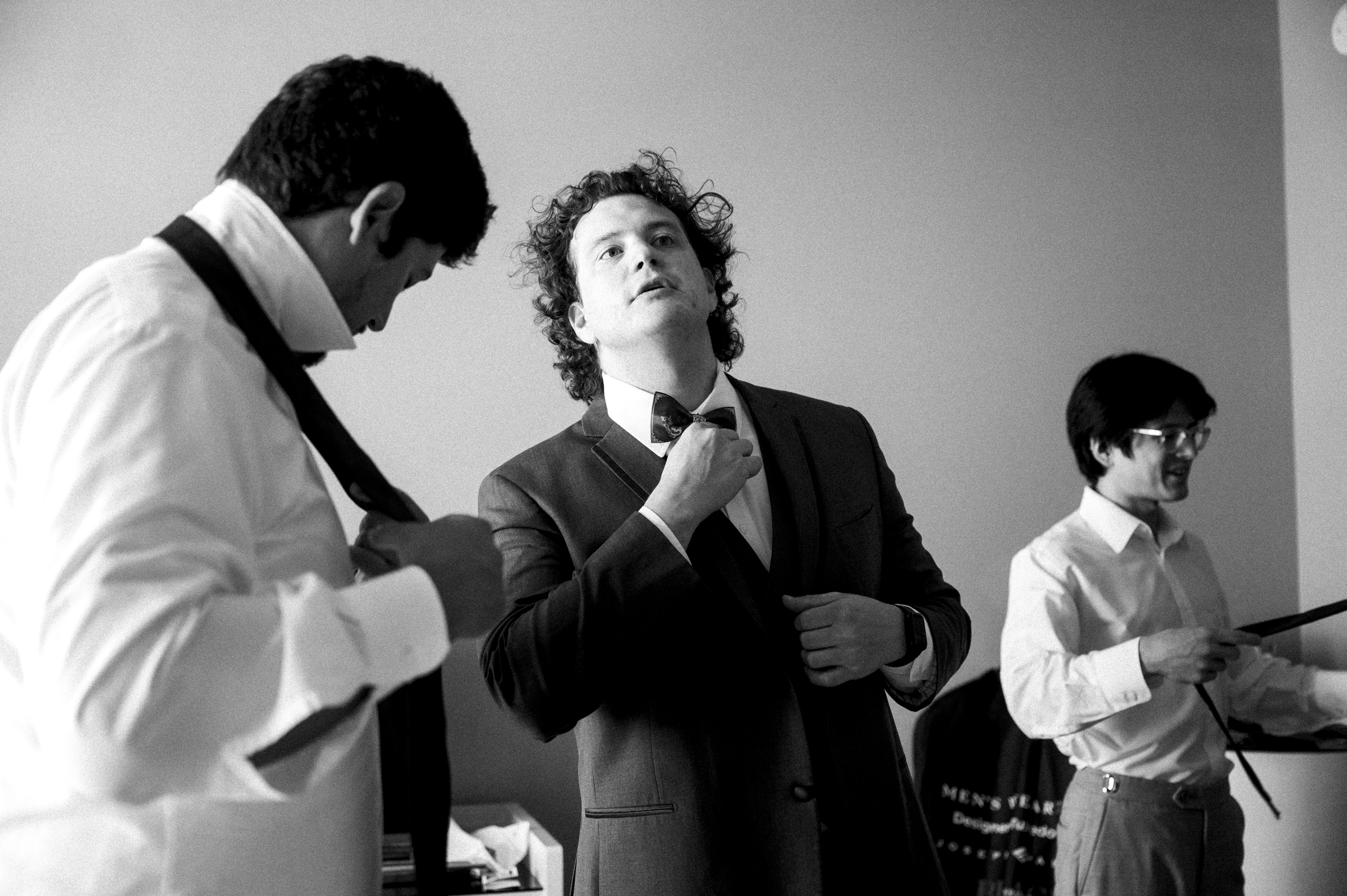 Three men in formal attire prepare for a Duke Chapel wedding. Two adjust their ties while the third stands confidently in a suit, looking at the camera. The atmosphere suggests anticipation and camaraderie, set against a simple indoor background.