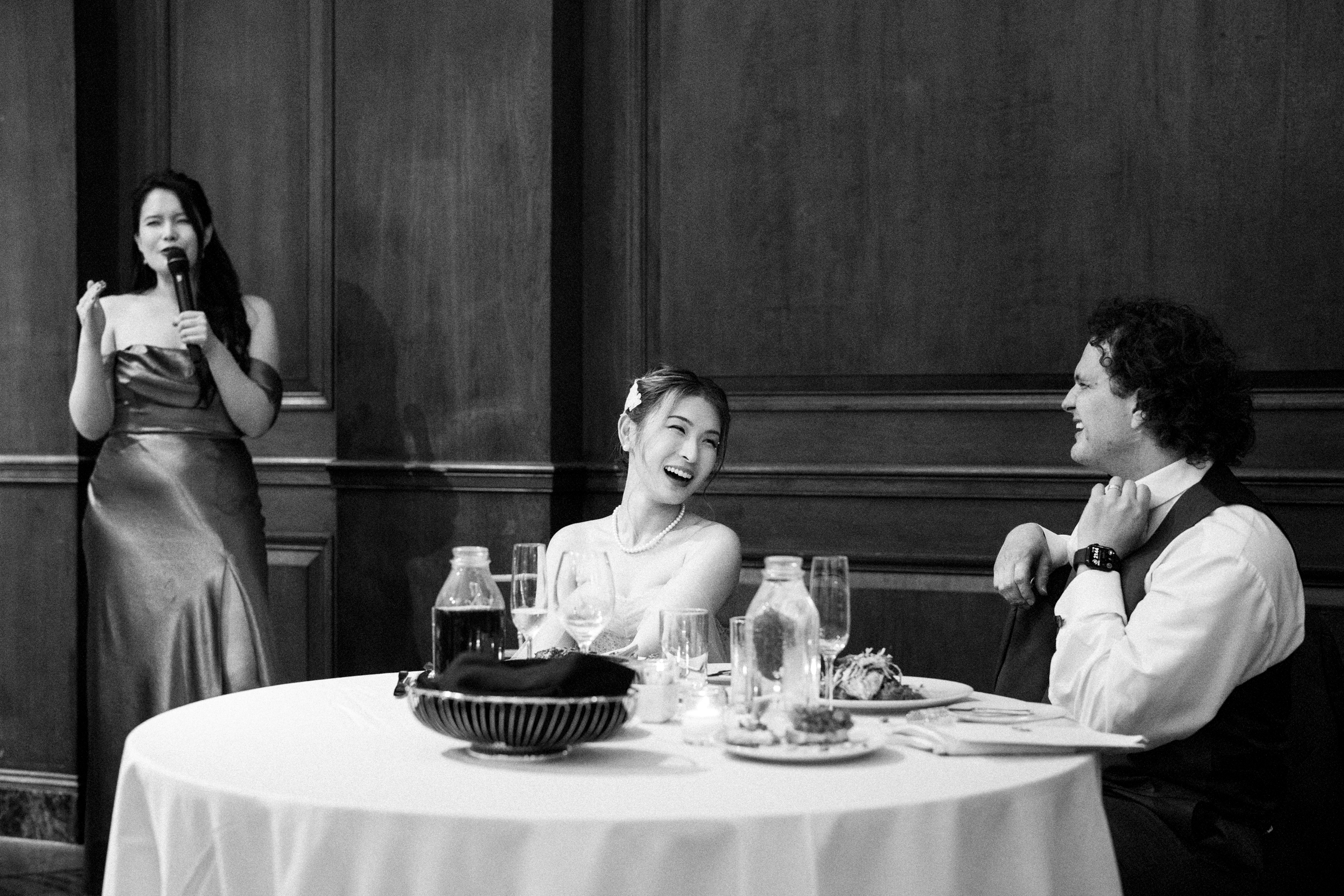 At a Duke Chapel wedding reception, a woman in a dress speaks into a microphone beside a seated couple at the table. The woman at the table laughs warmly, looking at the man who is smiling in the elegant dining area with wood paneling.